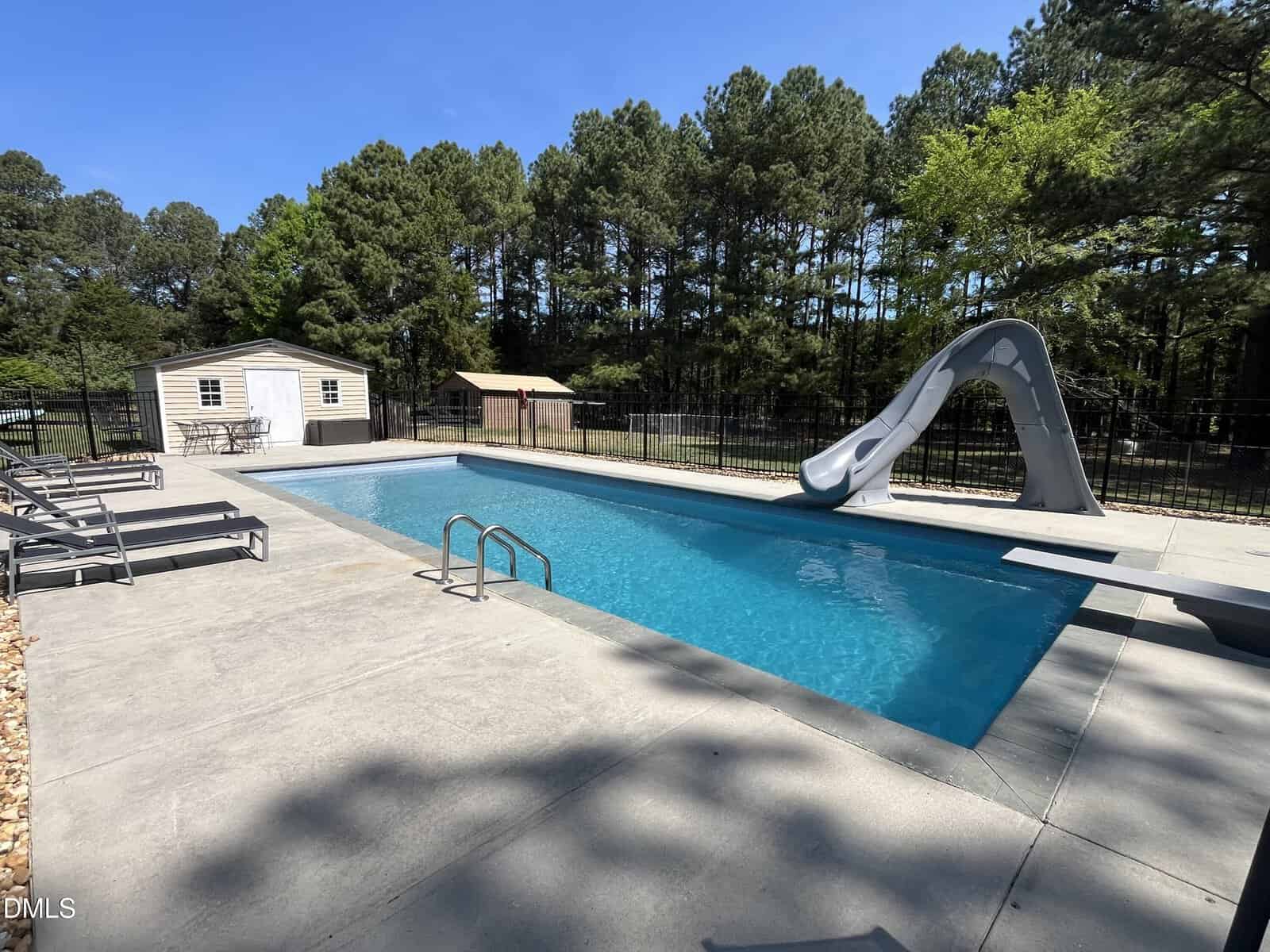 Rectangular outdoor swimming pool with a gray slide, metal handrails, and several lounge chairs on a concrete deck, surrounded by trees and a fence, with a small shed in the background.