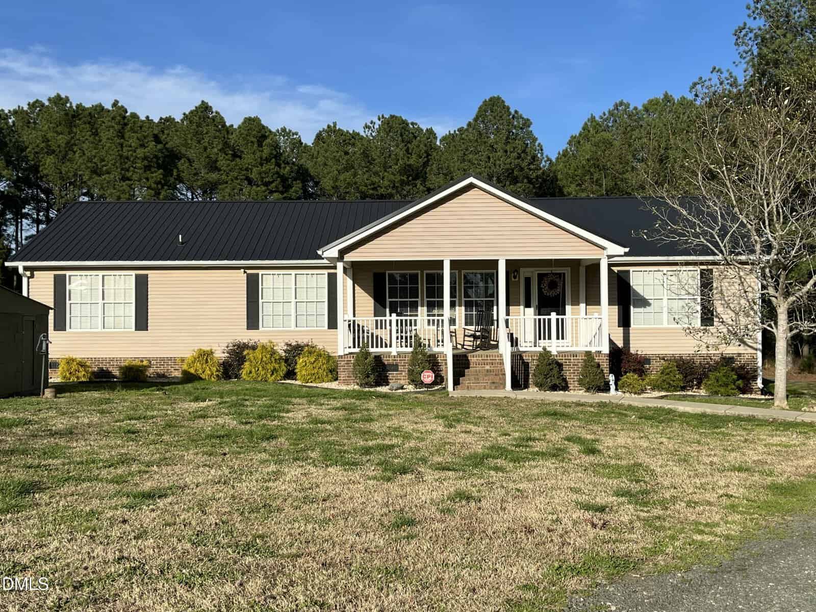 Nestled at 1195 US 158 in Oxford, this single-story beige house features a black roof, white trim, and a covered front porch with steps. The yard boasts patchy grass and small shrubs, bordered by tall pines under a blue sky.