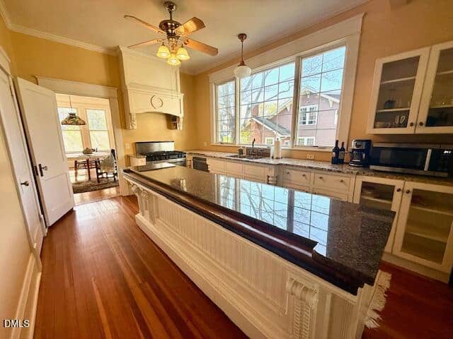 Bright kitchen with large island, granite countertop, white cabinets, stainless steel appliances, hardwood floors, and a ceiling fan. Big windows overlook a neighboring house. Dining area visible through open door.