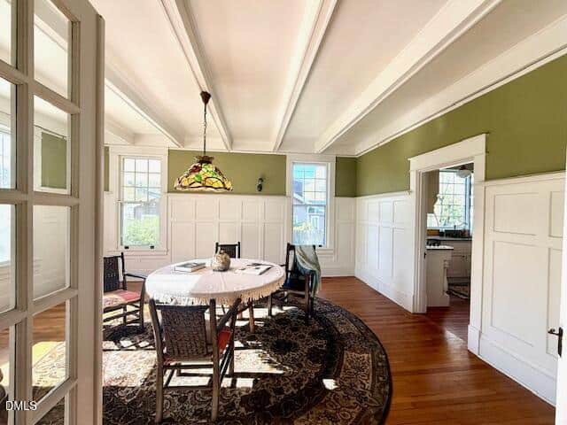 Sunny dining room with green and white walls, a round table with a covering, four chairs, stained glass pendant light, large windows, hardwood floors, and doorway leading to another room.