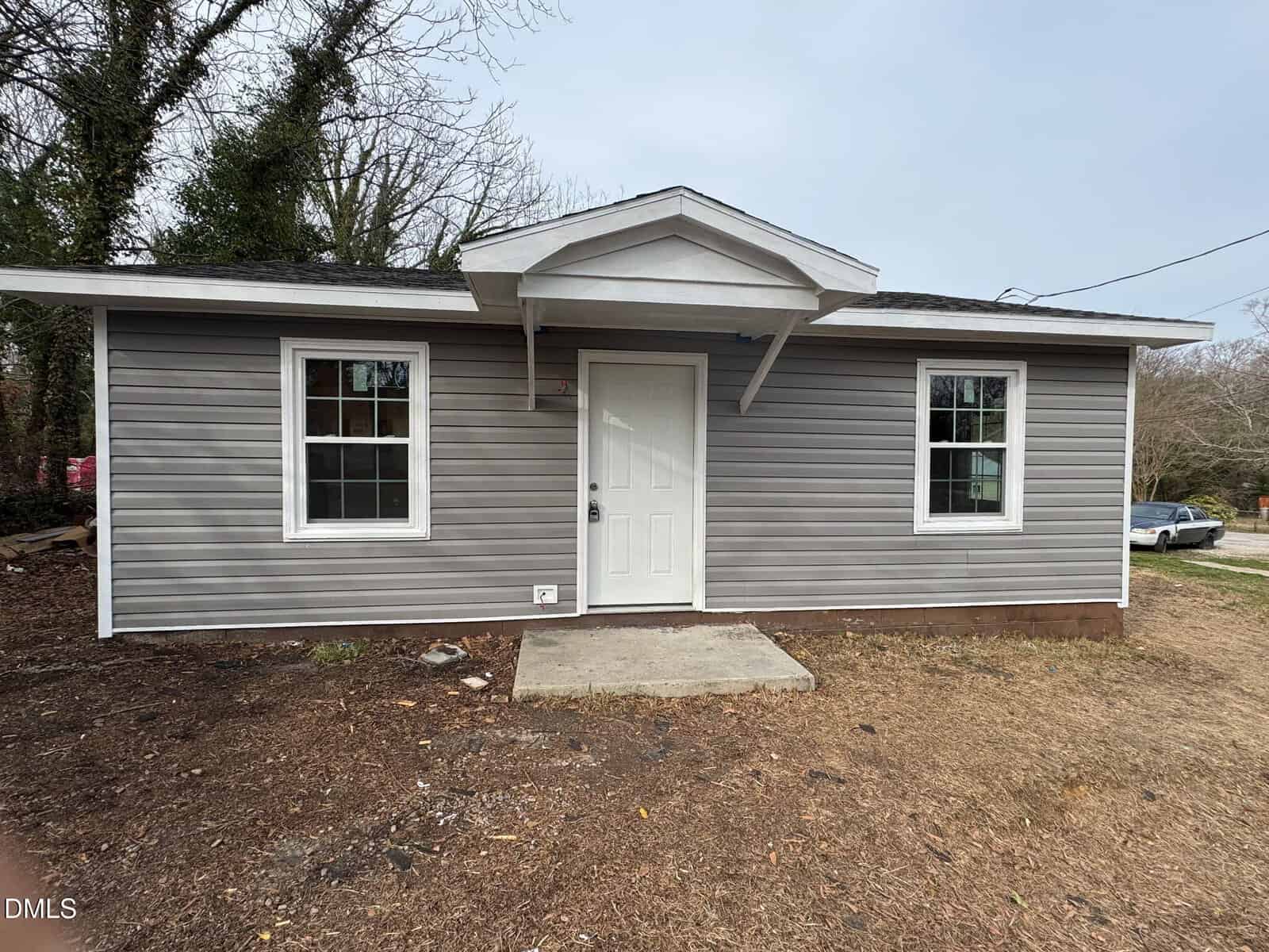 A small single-story house with gray vinyl siding, a white front door, two windows, and a small covered entryway. The yard is mostly bare dirt with patches of grass. Trees are visible in the background.