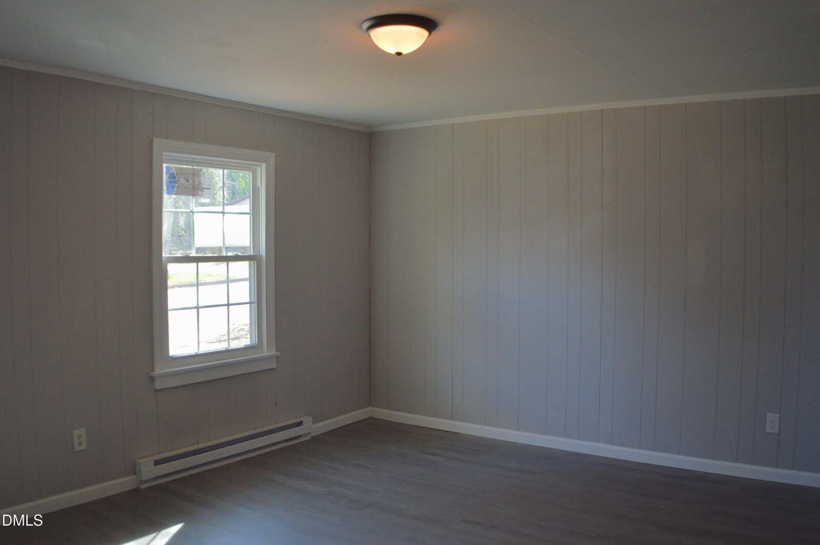 Empty room with gray paneled walls, a white-trimmed window letting in daylight, gray wood-look flooring, a ceiling light fixture, and a white baseboard heater under the window.