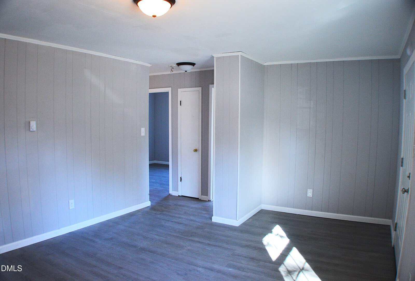 An empty room with light gray paneled walls, wood-look flooring, a white ceiling with a light fixture, and three doors leading to other areas. Sunlight is coming in through a window, casting shadows on the floor.