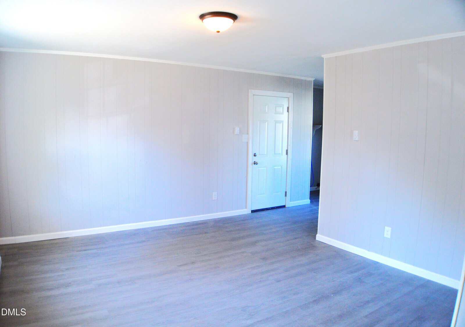 A room with light gray wood panel walls, gray wood flooring, a white ceiling, ceiling light fixture, and a white door. An alcove or entry area is visible to the right of the door.