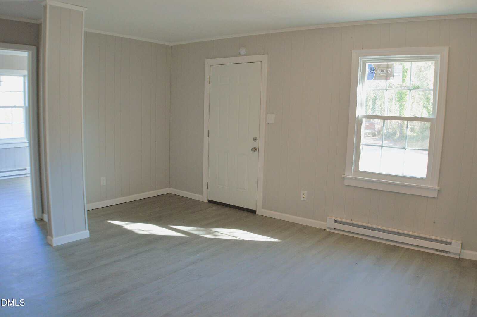A room with light gray paneled walls, light wood flooring, a white door, a window with sunlight streaming in, and a white baseboard heater below the window. An open doorway leads to another room.