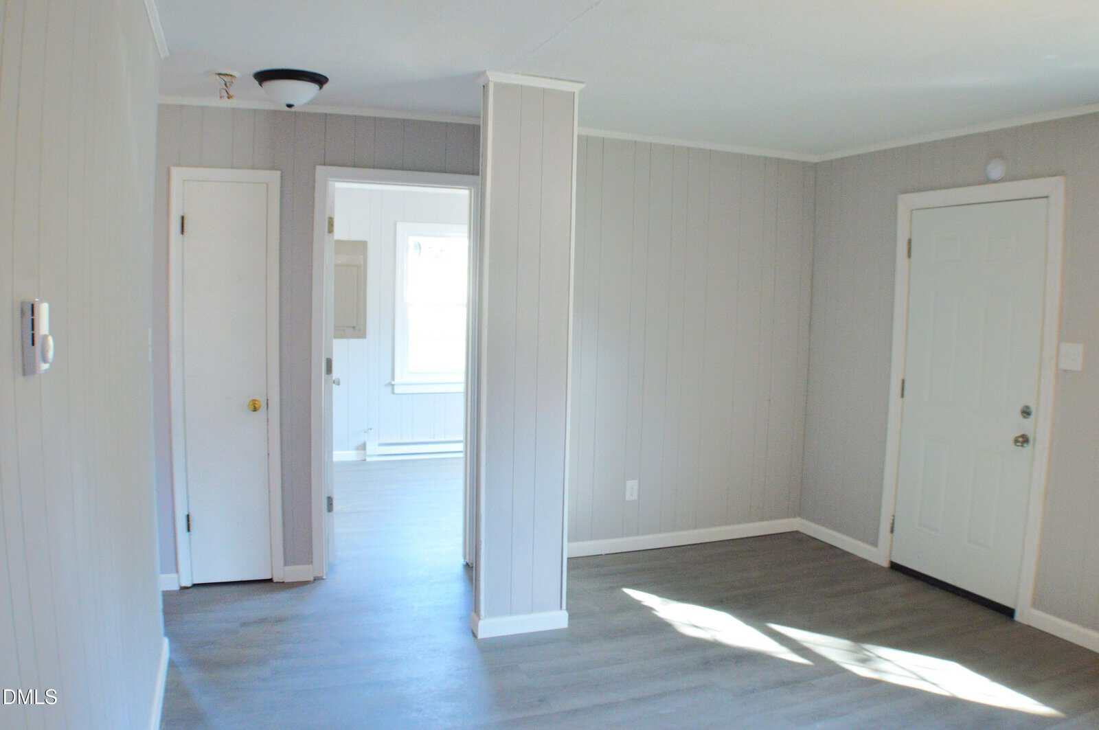 A small, empty room with light gray paneled walls, white trim, light wood flooring, a white door on the right, a small closet door on the left, and a partial wall dividing the space. Natural light enters through a window in the back.
