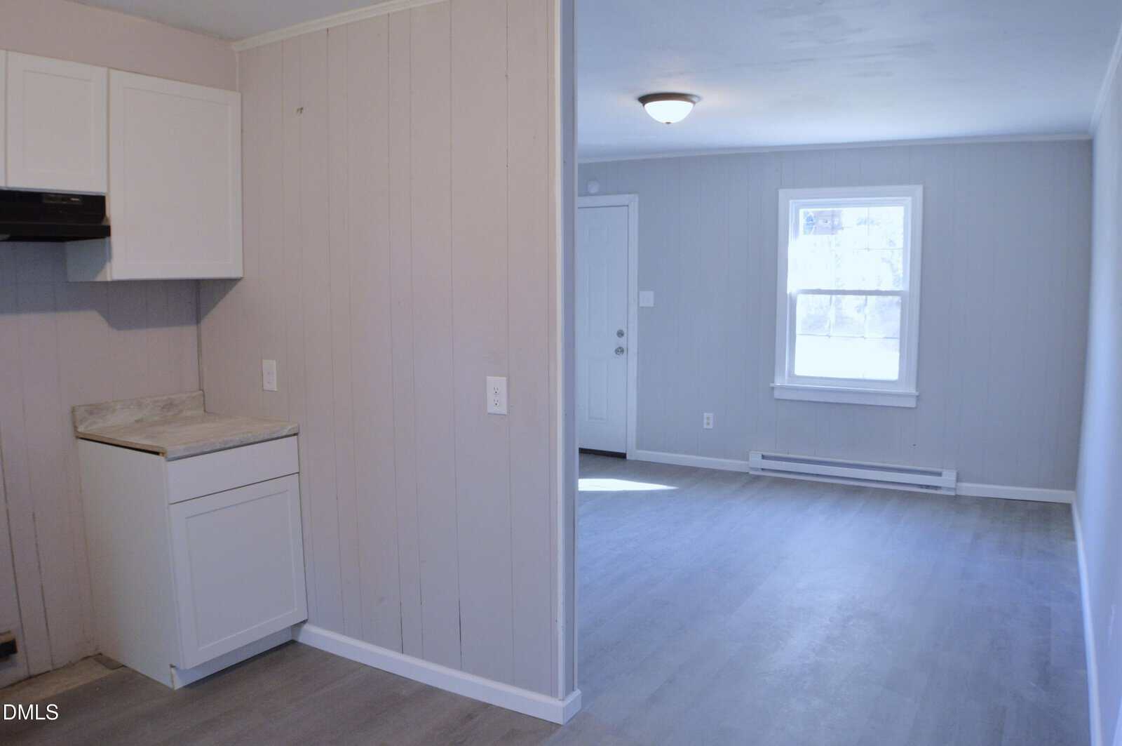 A small kitchen with light cabinets and marble countertop opens to a bright living area with a window, door, and light gray walls and flooring.