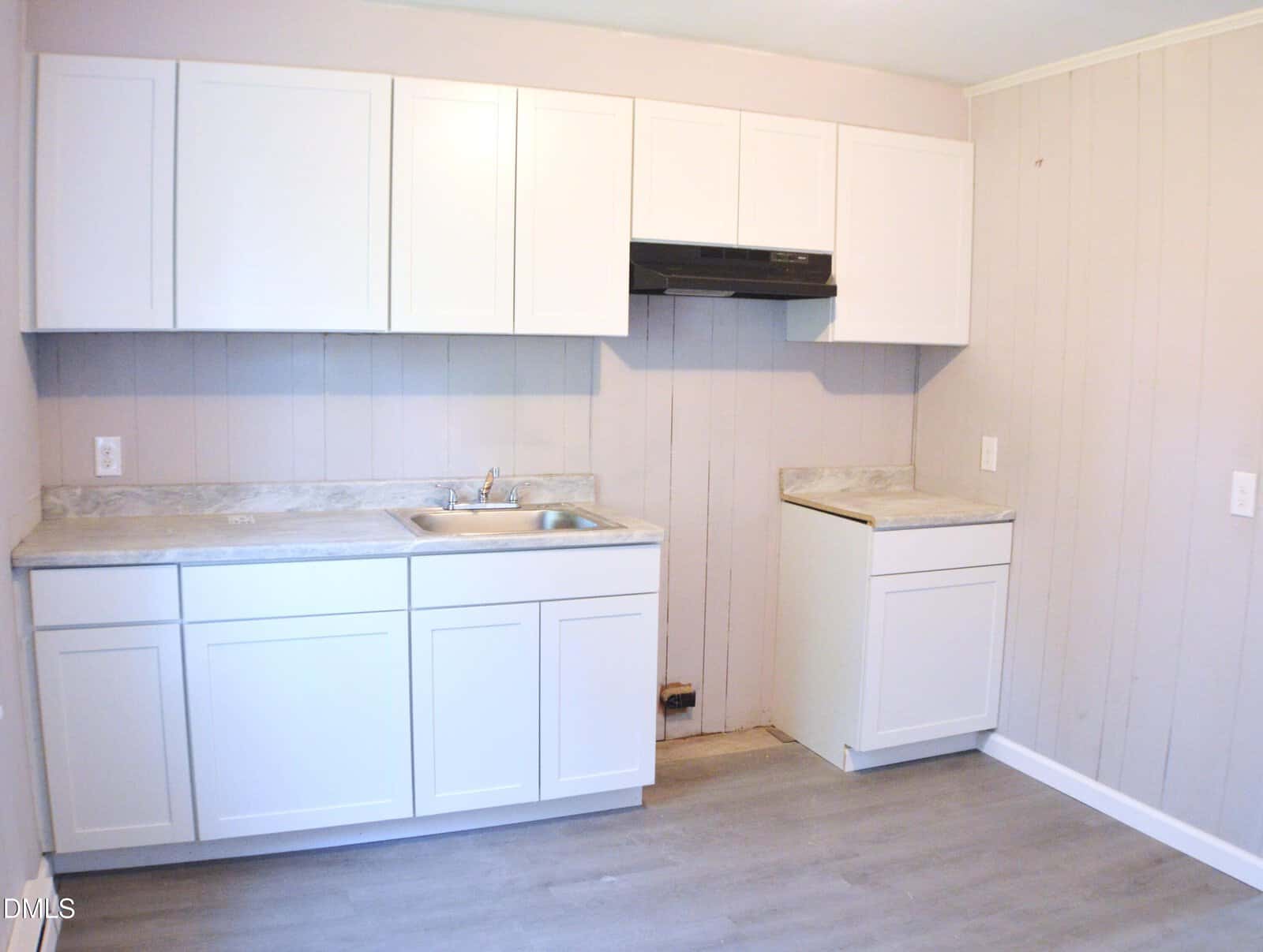 Small kitchen with white cabinets, marble-look countertops, a sink, a range hood, and light wood panel walls. There’s an empty space for a stove or appliance beside the right cabinet.