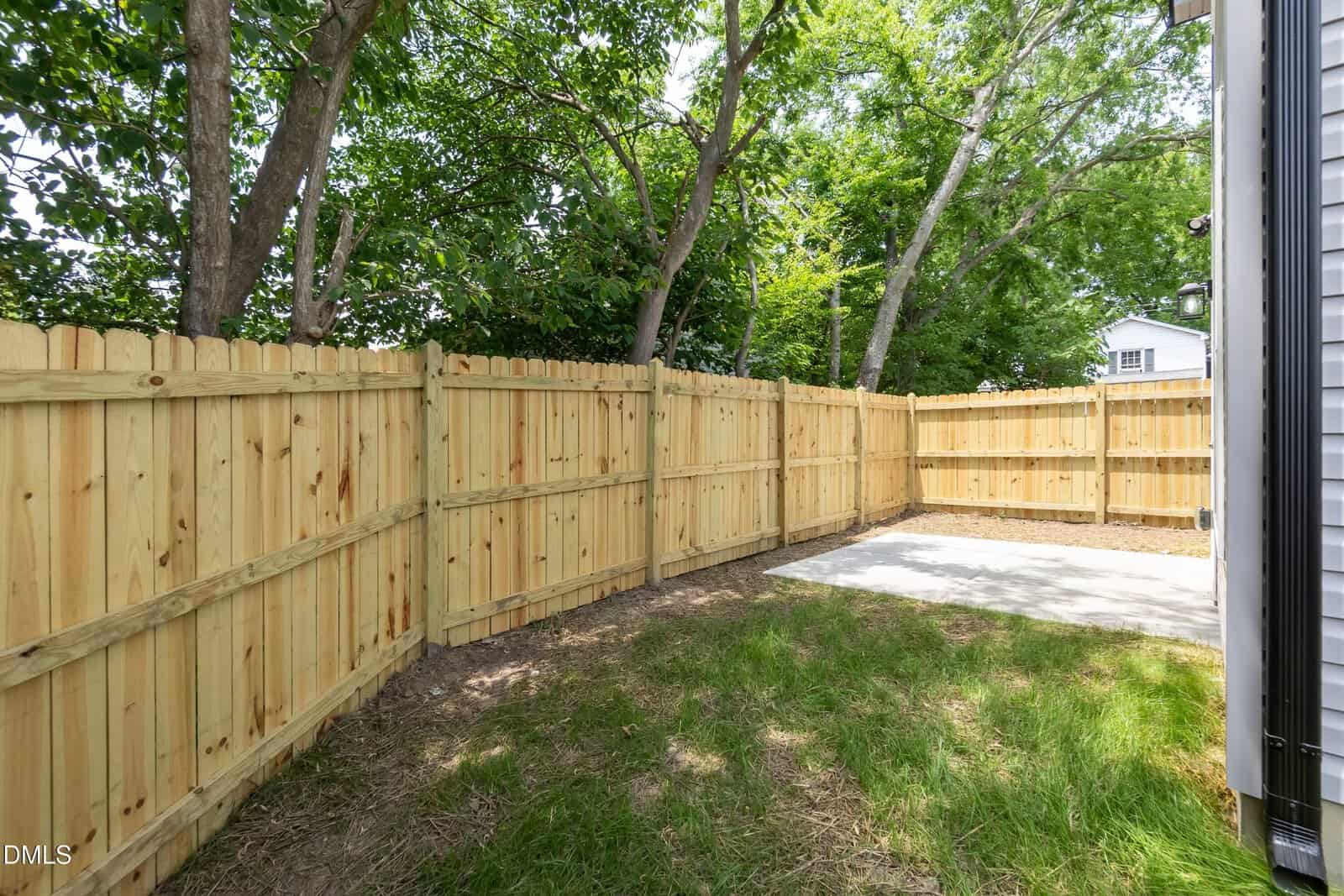A backyard with a wooden privacy fence, green grass, trees in the background, and a small concrete patio next to the house.