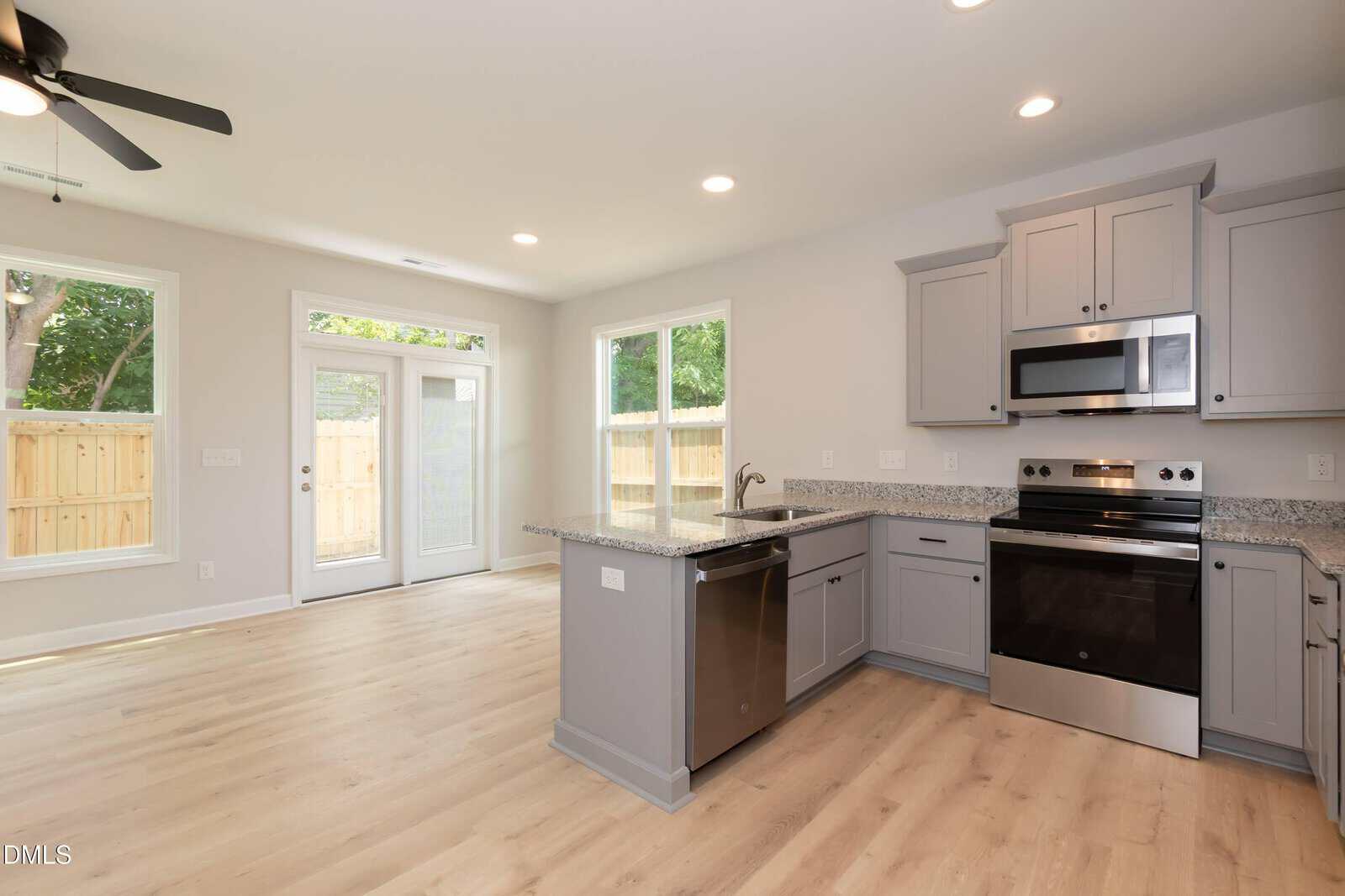 Modern kitchen with gray cabinets, stainless steel appliances, and light wood flooring. Large windows and glass doors provide ample natural light and view of a fenced backyard. Ceiling fan is visible in the adjacent dining area.