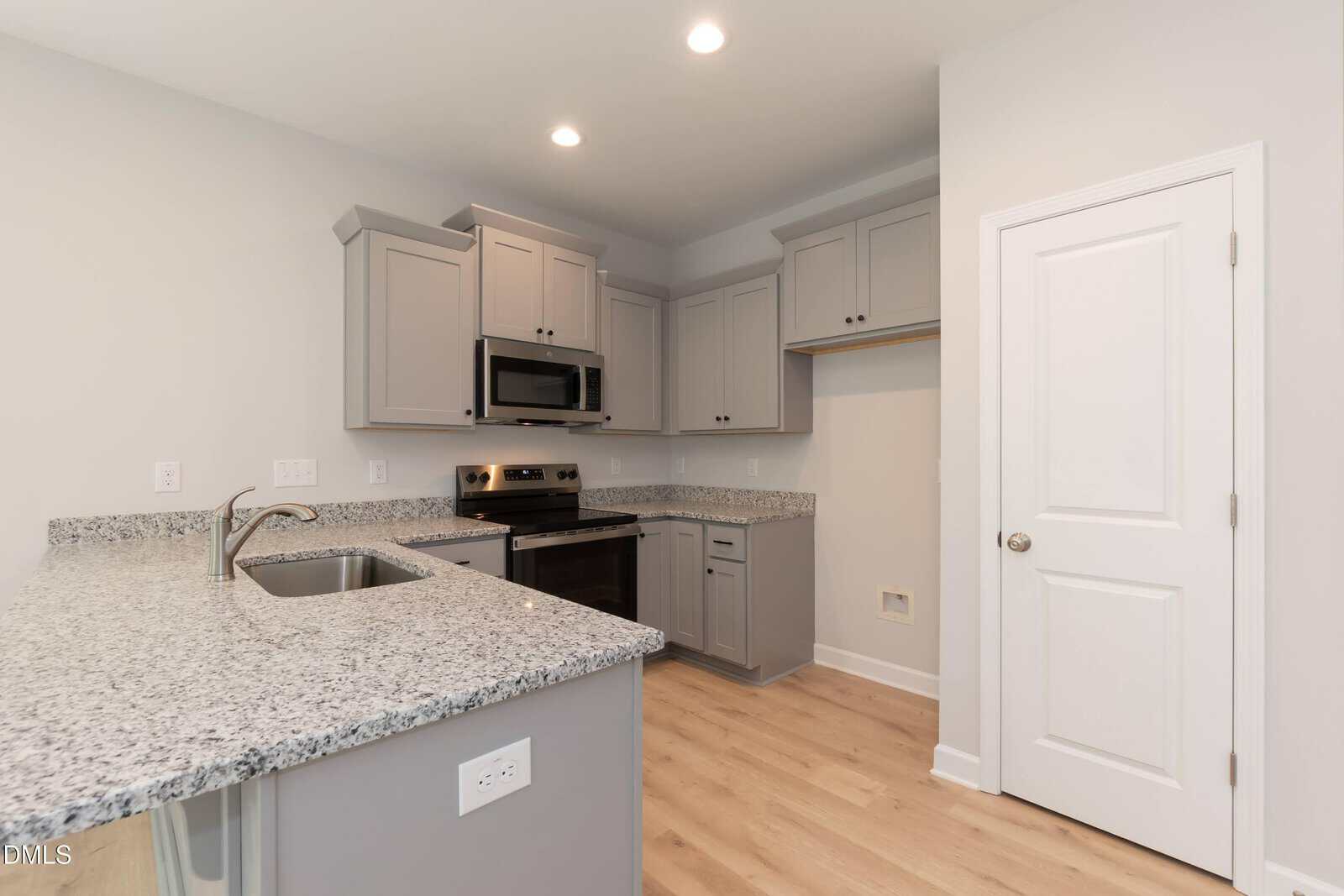 Modern kitchen with light gray cabinets, granite countertops, stainless steel appliances, built-in microwave, electric stove, stainless steel sink, and light wood flooring. A white door and open pantry are also visible.