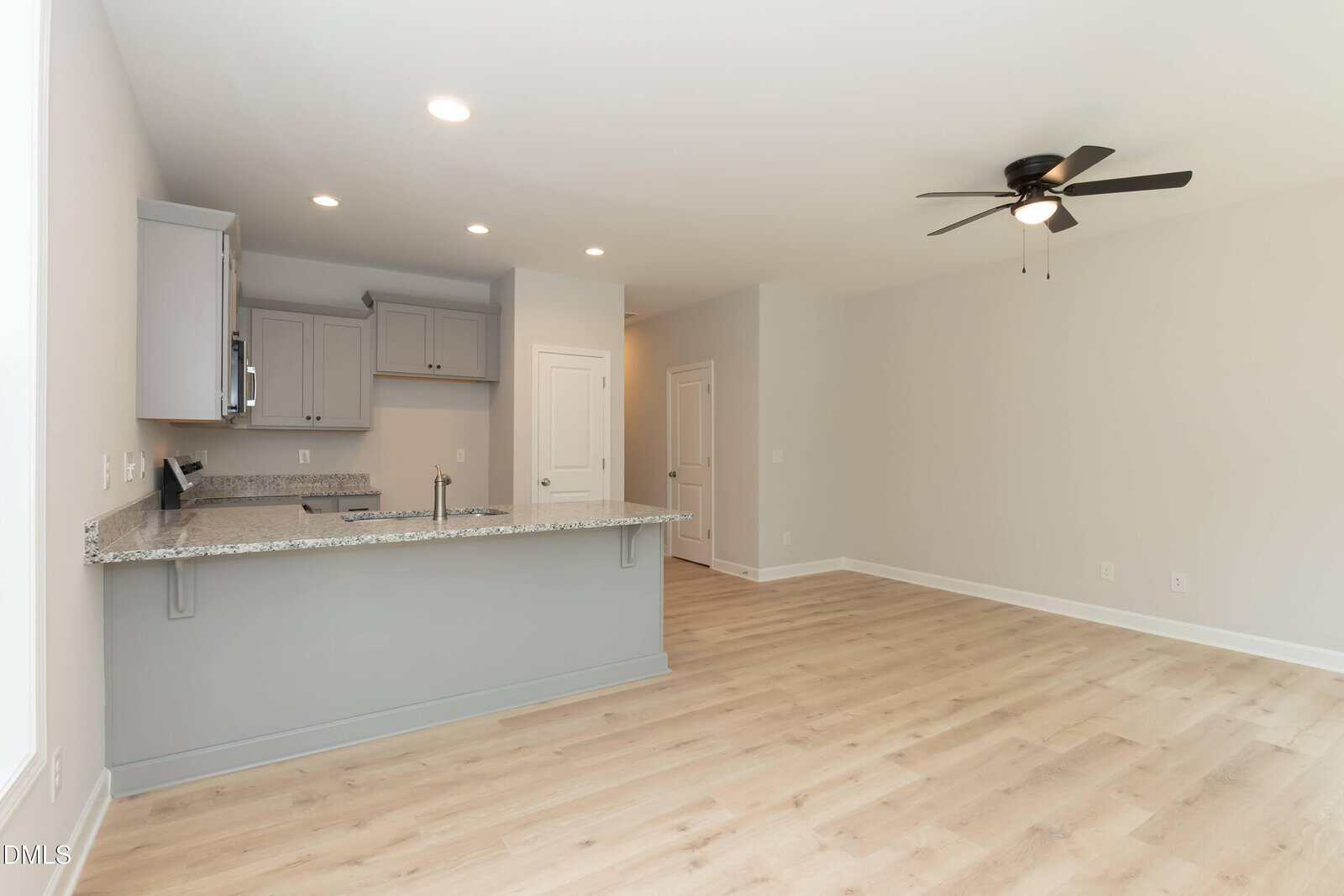 Modern, unfurnished open-plan kitchen and living area with light wood flooring, gray cabinets, granite countertops, recessed lighting, ceiling fan, and neutral-colored walls.