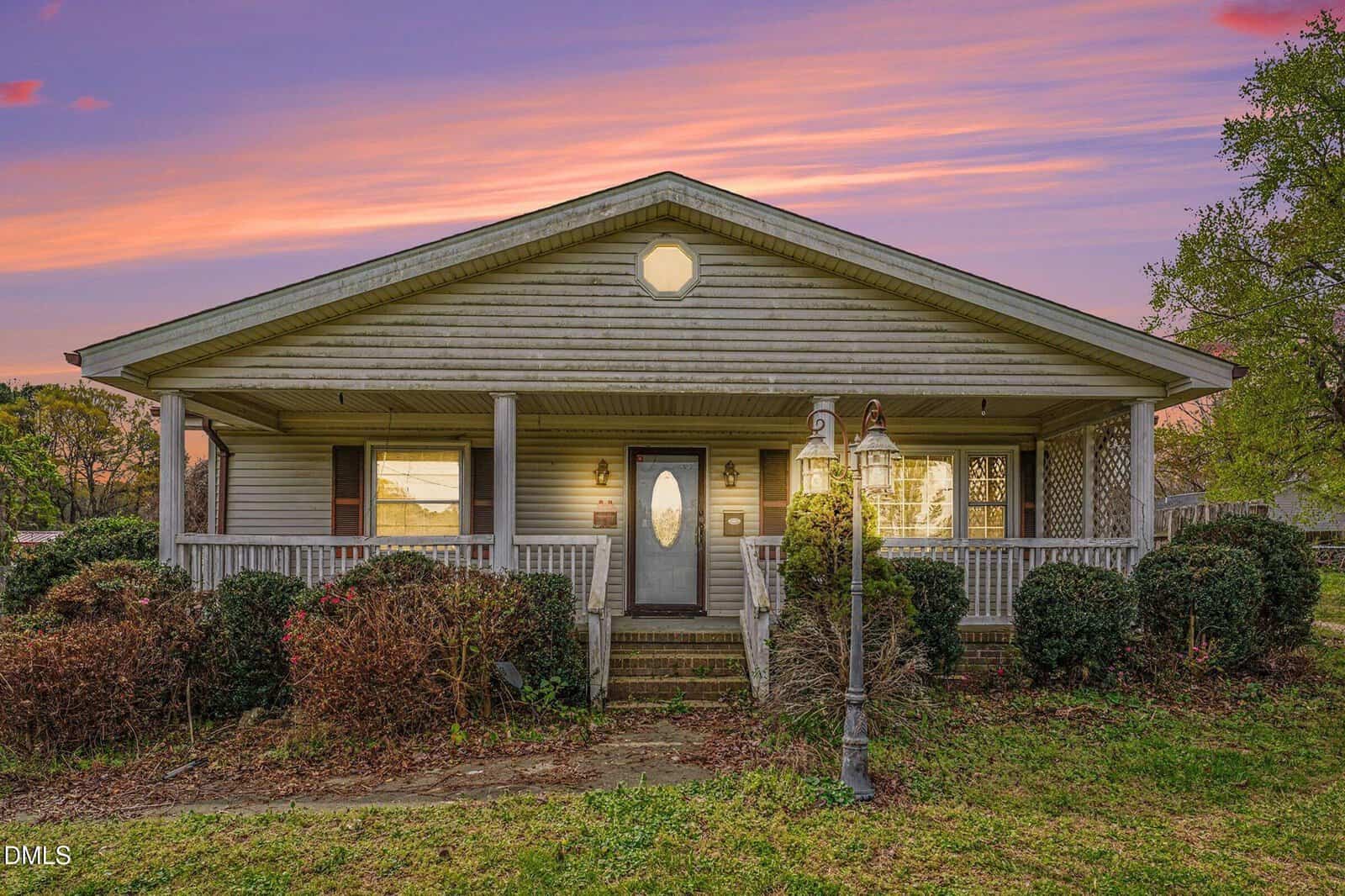 Single-story house with a covered front porch, central door with decorative glass, and surrounding bushes. The sky is colorful at sunset, with a double lamp post in the yard.