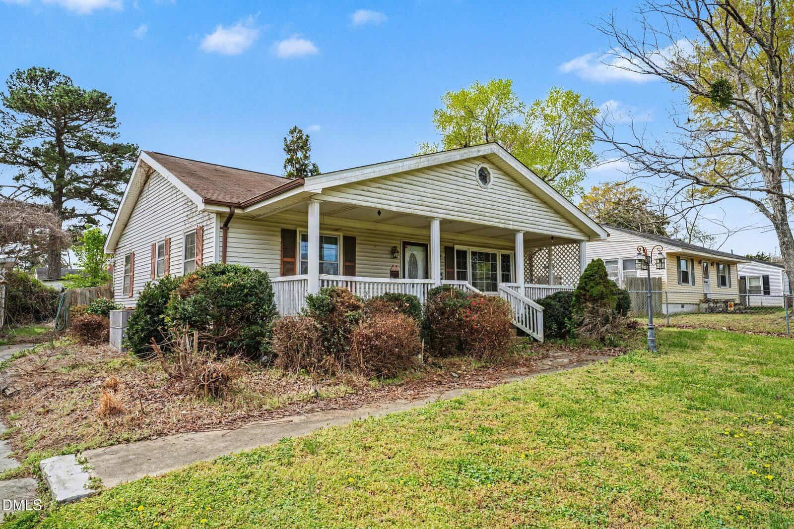 Single-story house with beige siding and a covered front porch, surrounded by bushes and a grassy yard. A concrete walkway leads to the porch, and another house is visible to the right. The sky is clear with a few clouds.