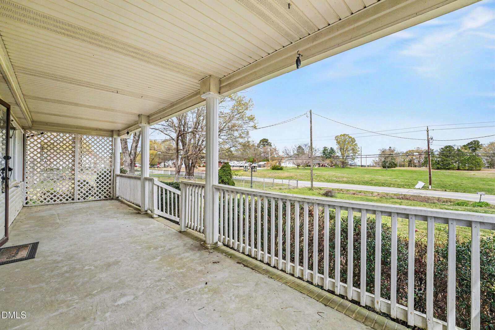 A covered front porch with a white railing and concrete floor overlooks a grassy lawn and a road lined with trees and utility poles under a partly cloudy sky.
