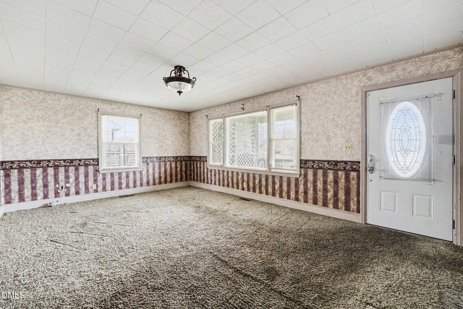 Empty living room with patterned wallpaper, green carpet, three windows, a white front door with decorative glass, and a ceiling light fixture. Natural light enters from the windows.