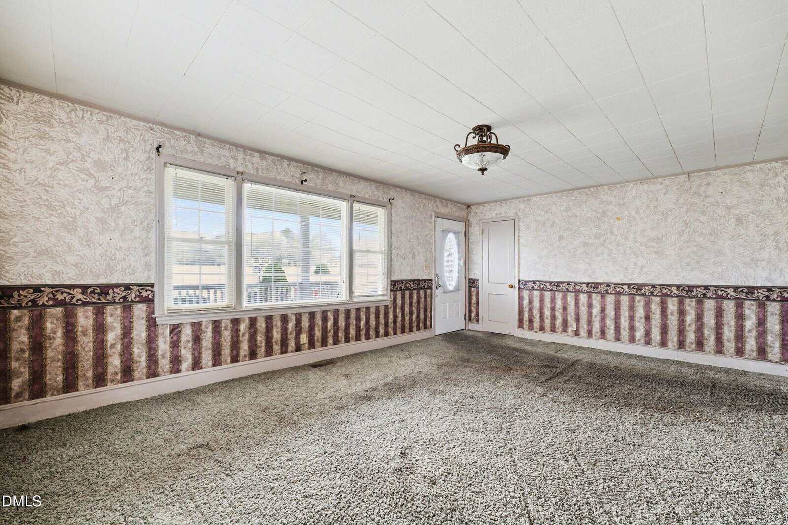 Empty room with patterned wallpaper, worn carpet, three large windows, a white door with an oval window, and a vintage ceiling light fixture. Walls have a textured look with a decorative border.