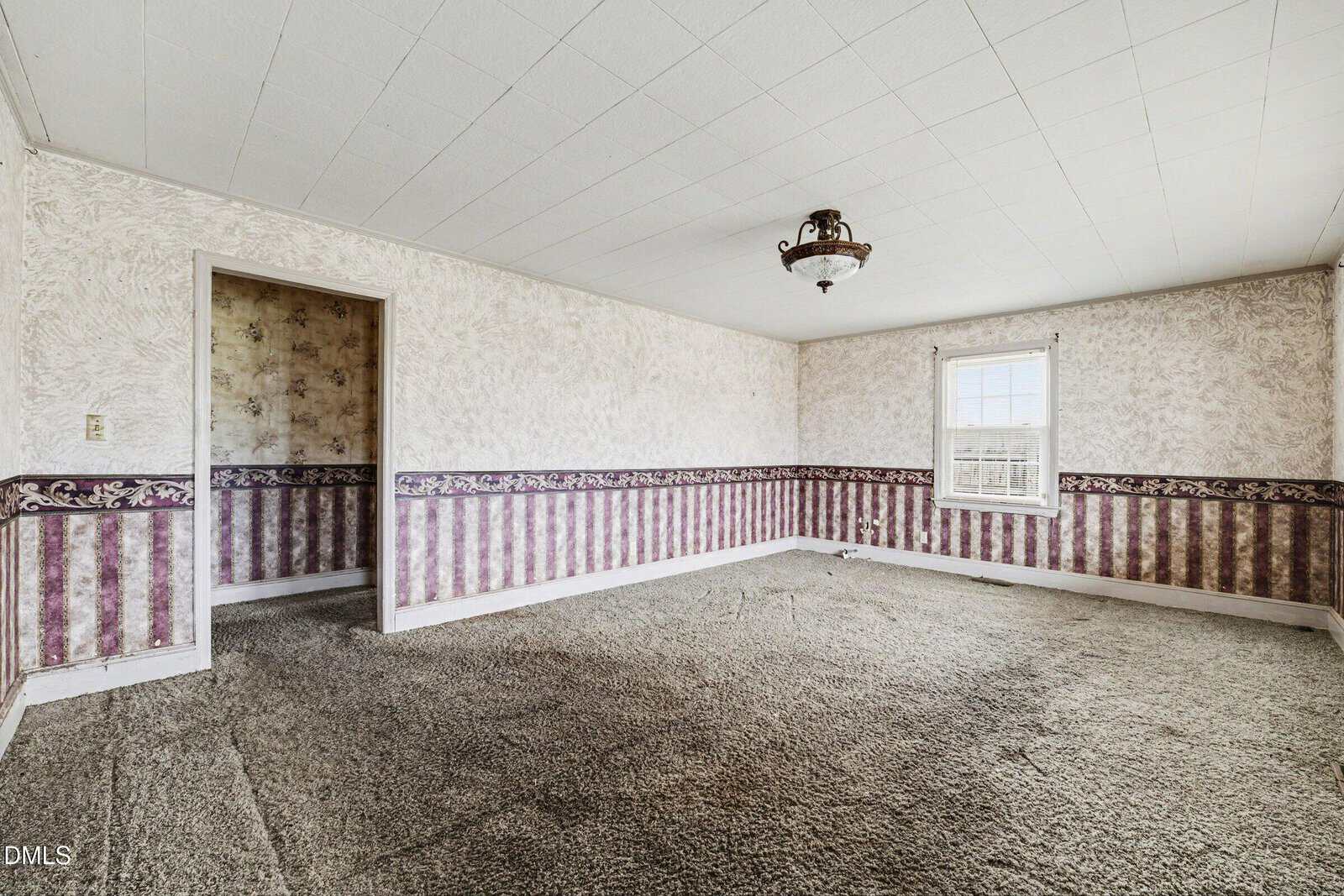 Empty room with patterned wallpaper, old carpet, a single window letting in light, and a vintage ceiling light fixture. Doorway leads to another room with similar decor.