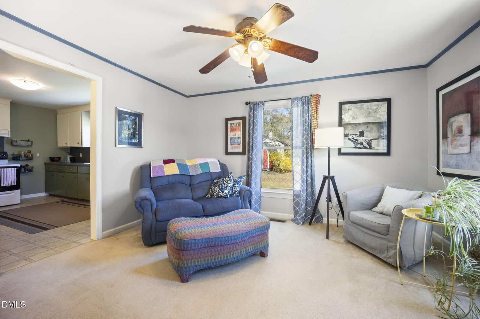 A living room with a blue recliner, a round ottoman, a gray armchair, a floor lamp, framed artwork on the walls, and a ceiling fan with lights. A window with blue curtains shows a view of the yard outside.
