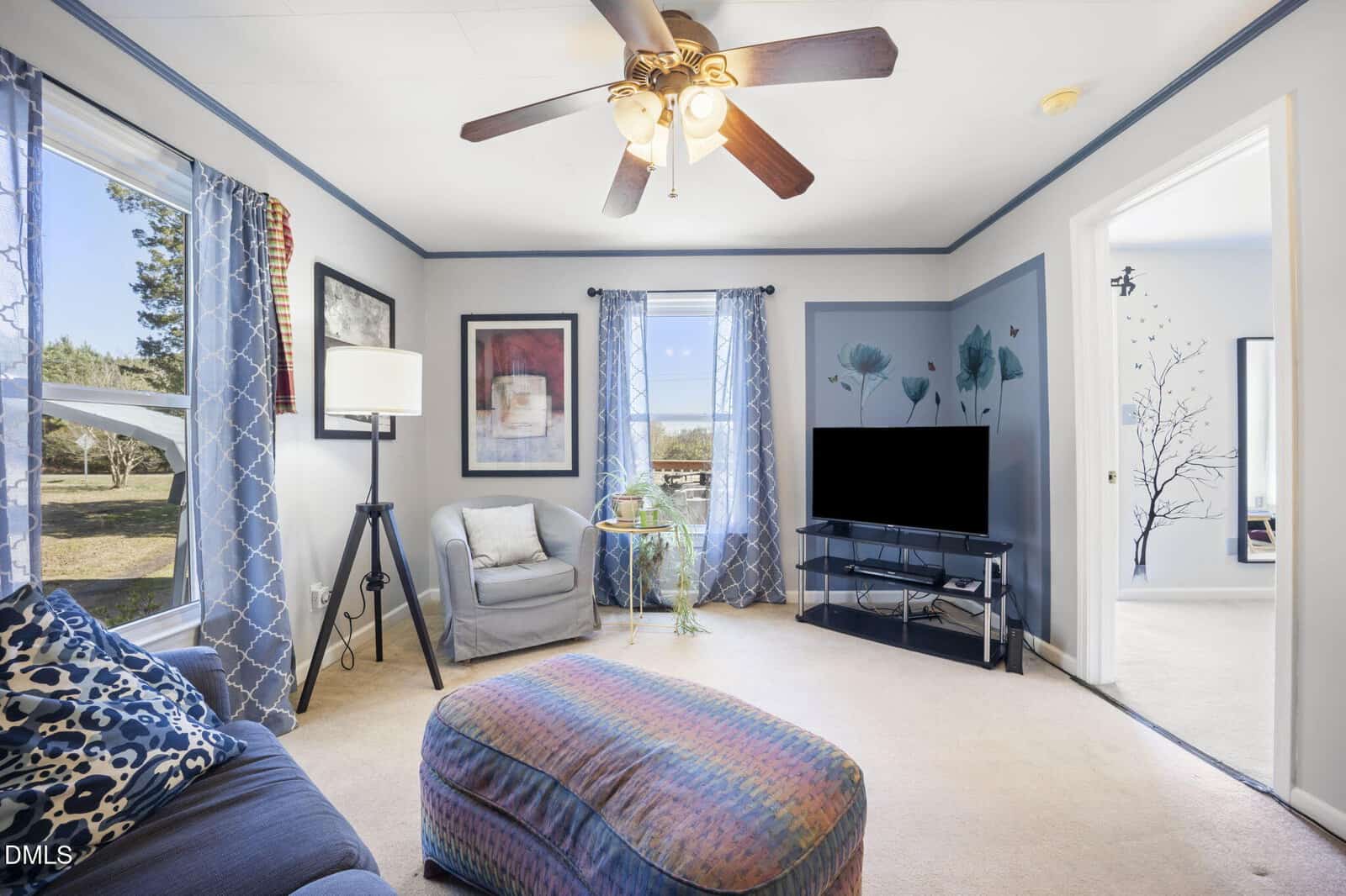 A living room with light-colored carpet, blue and gray furniture, a patterned ottoman, a TV on a glass stand, art on the walls, a ceiling fan, and two windows with blue curtains letting in natural light.