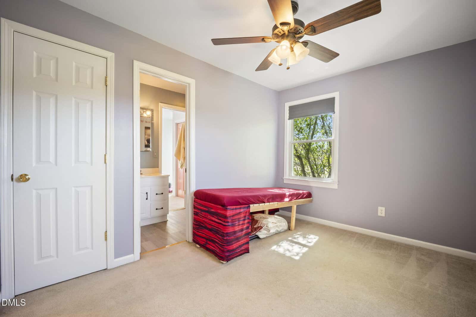 A simple bedroom with beige carpet, pale purple walls, a ceiling fan, a window with a tree view, a closed closet door, and a red massage table near an open door to a bathroom.