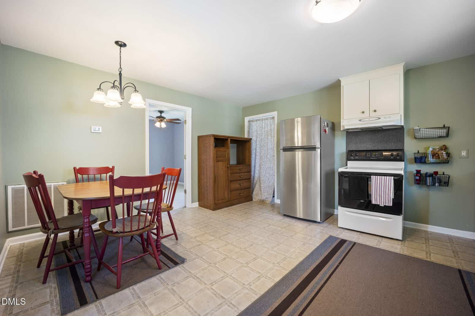 A kitchen with a dining area featuring a wooden table and red chairs, a stainless steel refrigerator, a white oven with range hood, wooden shelving, pale green walls, and a tile-patterned floor.