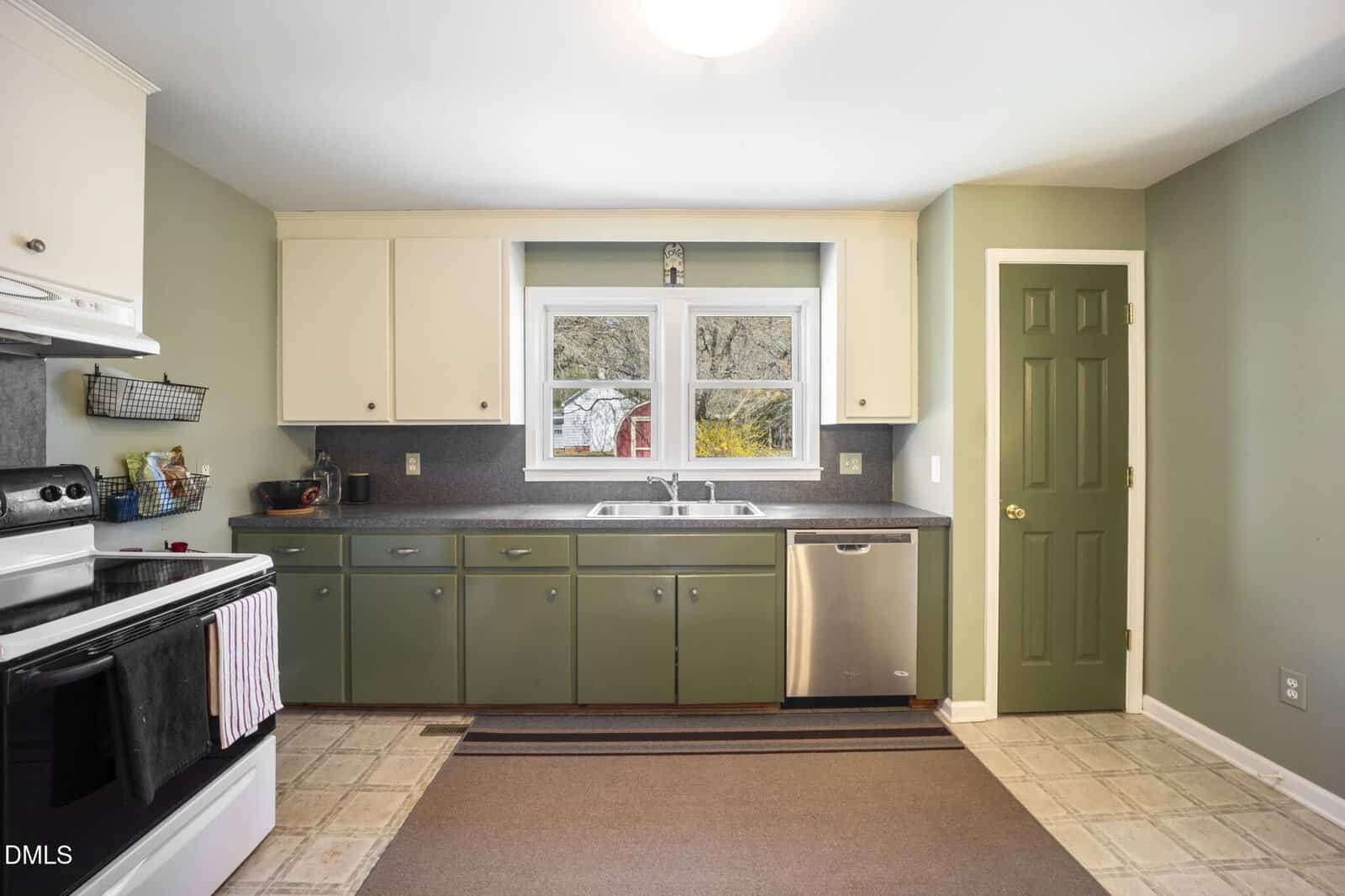 A kitchen with green lower cabinets, white upper cabinets, a stainless steel dishwasher, a double sink under a window, and a green door. The floor is tiled, and a striped rug lies in front of the sink.