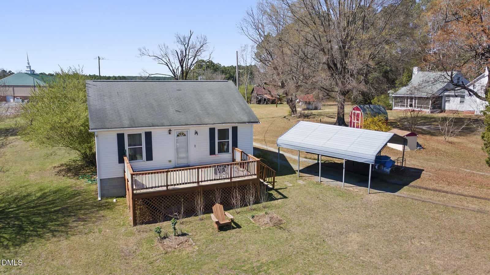 A small white house with a front porch and wooden steps sits on a grassy lot. Nearby, there is a covered carport, a red shed, and several mature trees. Neighboring homes and a clear sky are visible in the background.