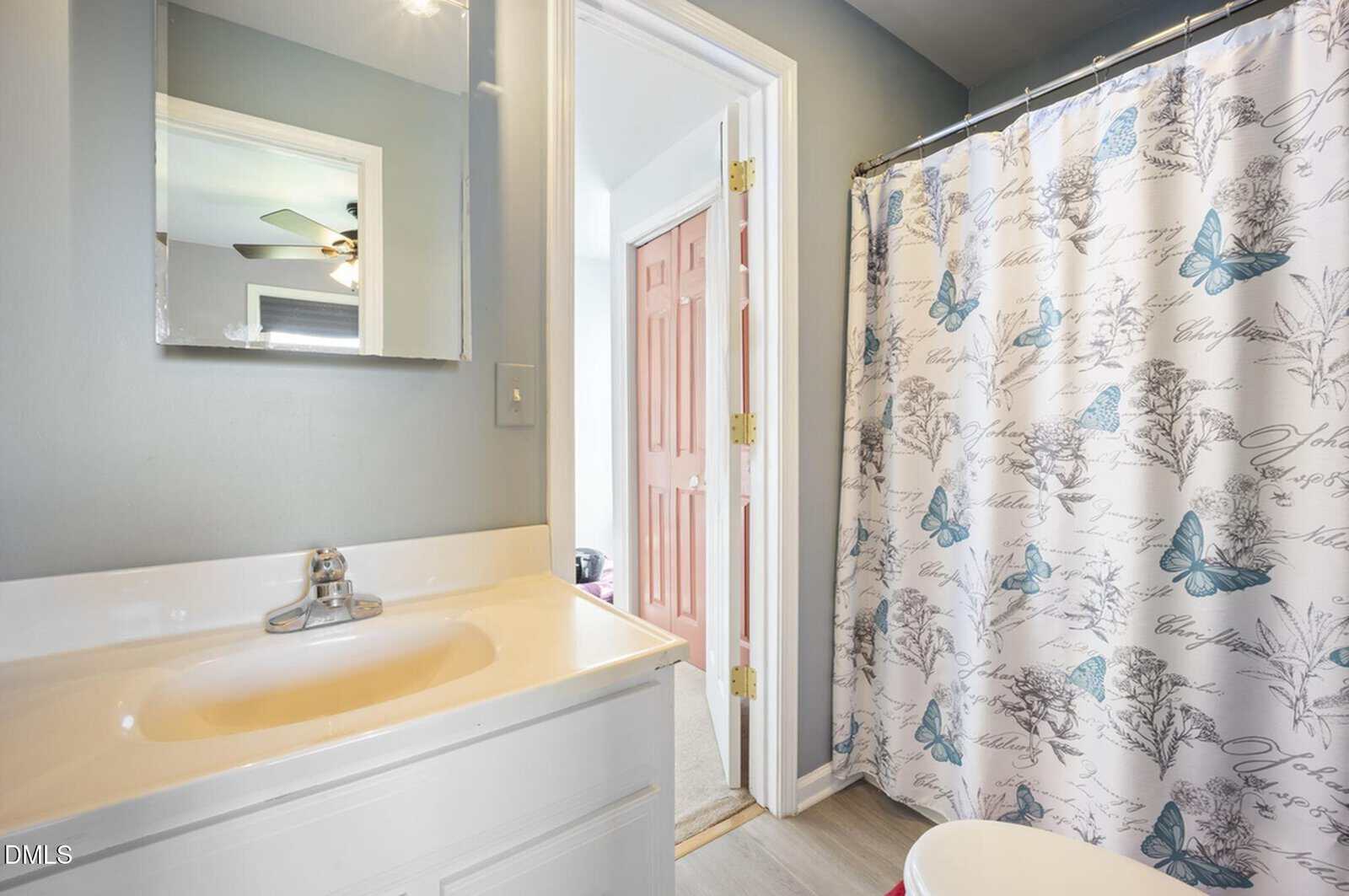 Bathroom with a white sink and cabinet, a mirror above, and a shower with a white curtain featuring blue butterflies and script. A door is open to an adjacent room with a reddish door.