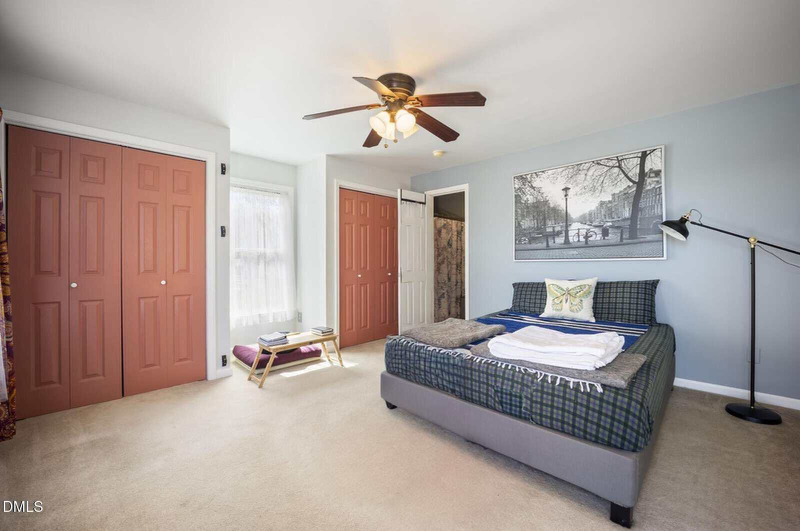 Bedroom with tan carpet, blue-gray walls, a bed with plaid bedding, artwork above, a floor lamp, ceiling fan, two large reddish closets, and a window with sheer curtains. A bench and books sit near the window.