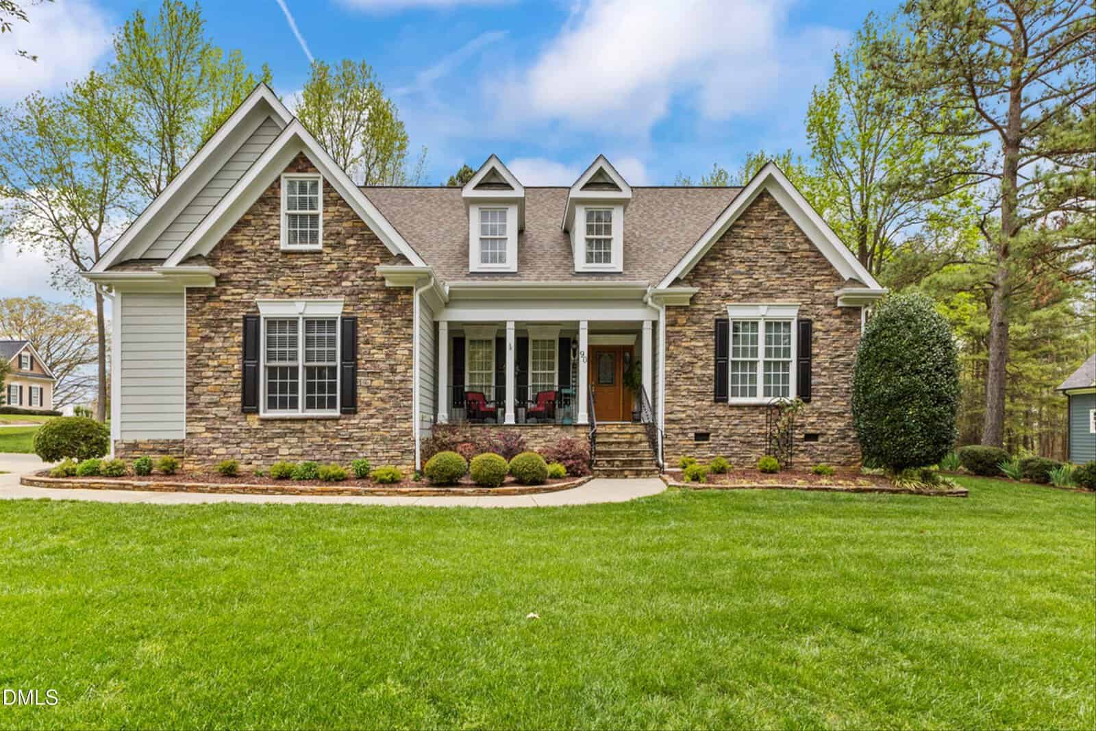 Exterior front view of a stone-facade house with multiple gabled dormers, black shutters, a covered front porch, landscaped lawn, and surrounding trees under a partly cloudy sky.