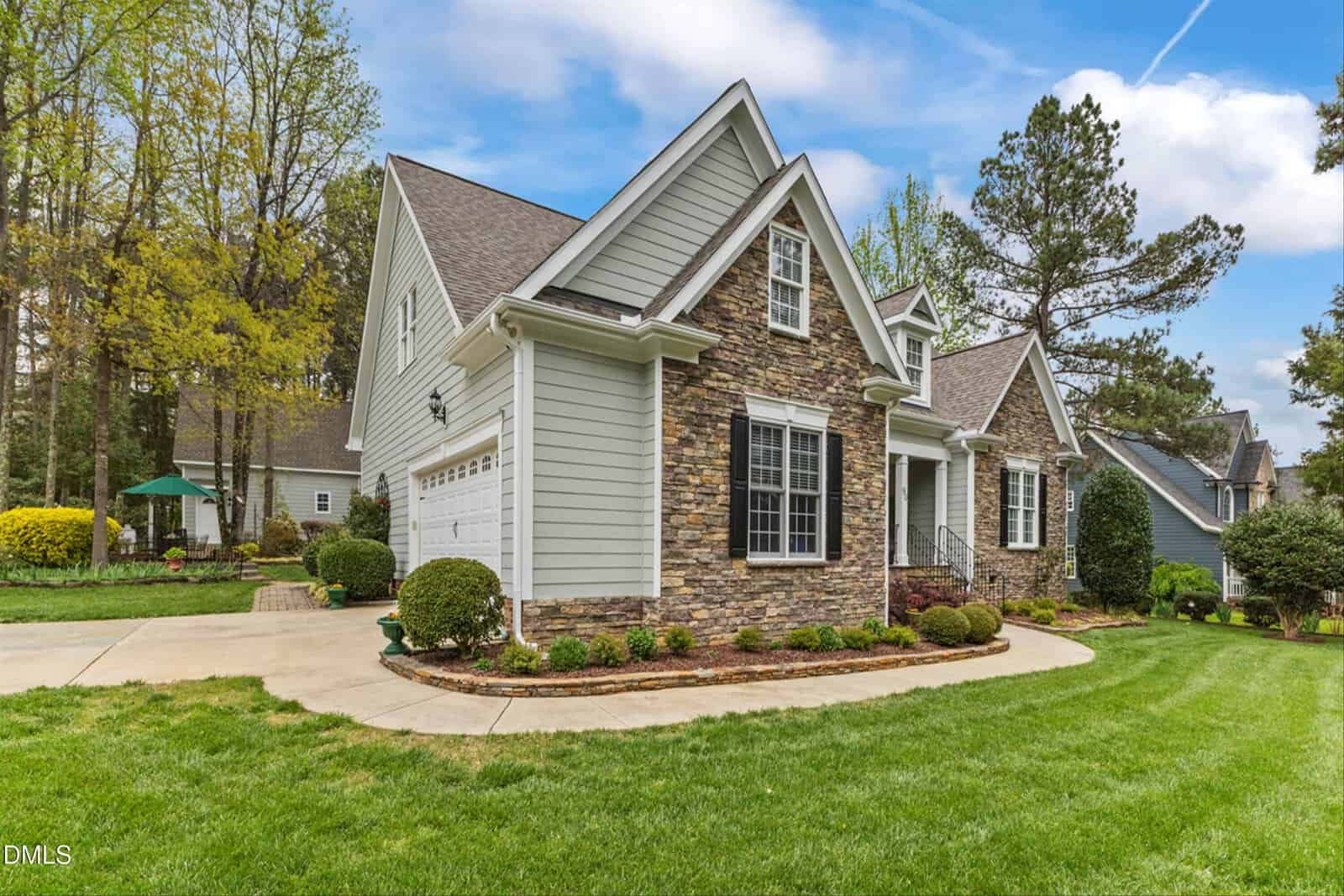A single-family house with gray siding, stone accents, white trim, and a two-car garage sits on a well-kept lawn with shrubs and a curved driveway, surrounded by trees and neighboring homes.