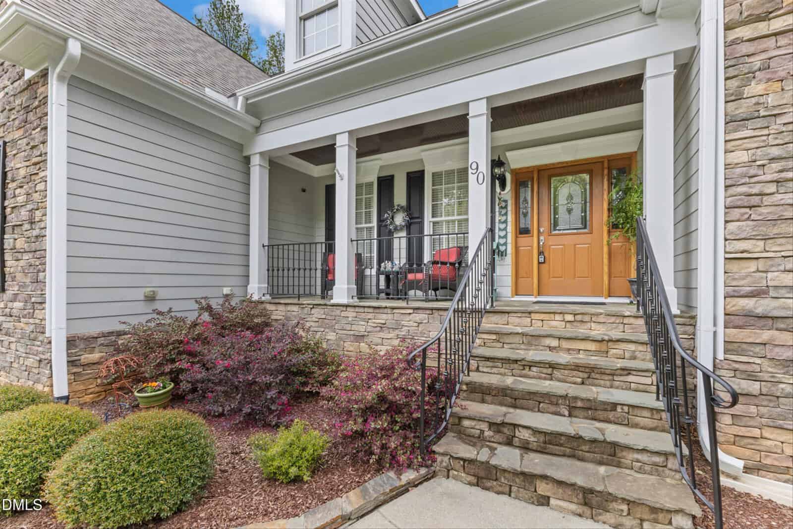 A stone staircase with black railings leads to a covered porch with white columns, gray siding, and a wooden front door. Two red-cushioned chairs and potted plants decorate the porch area.