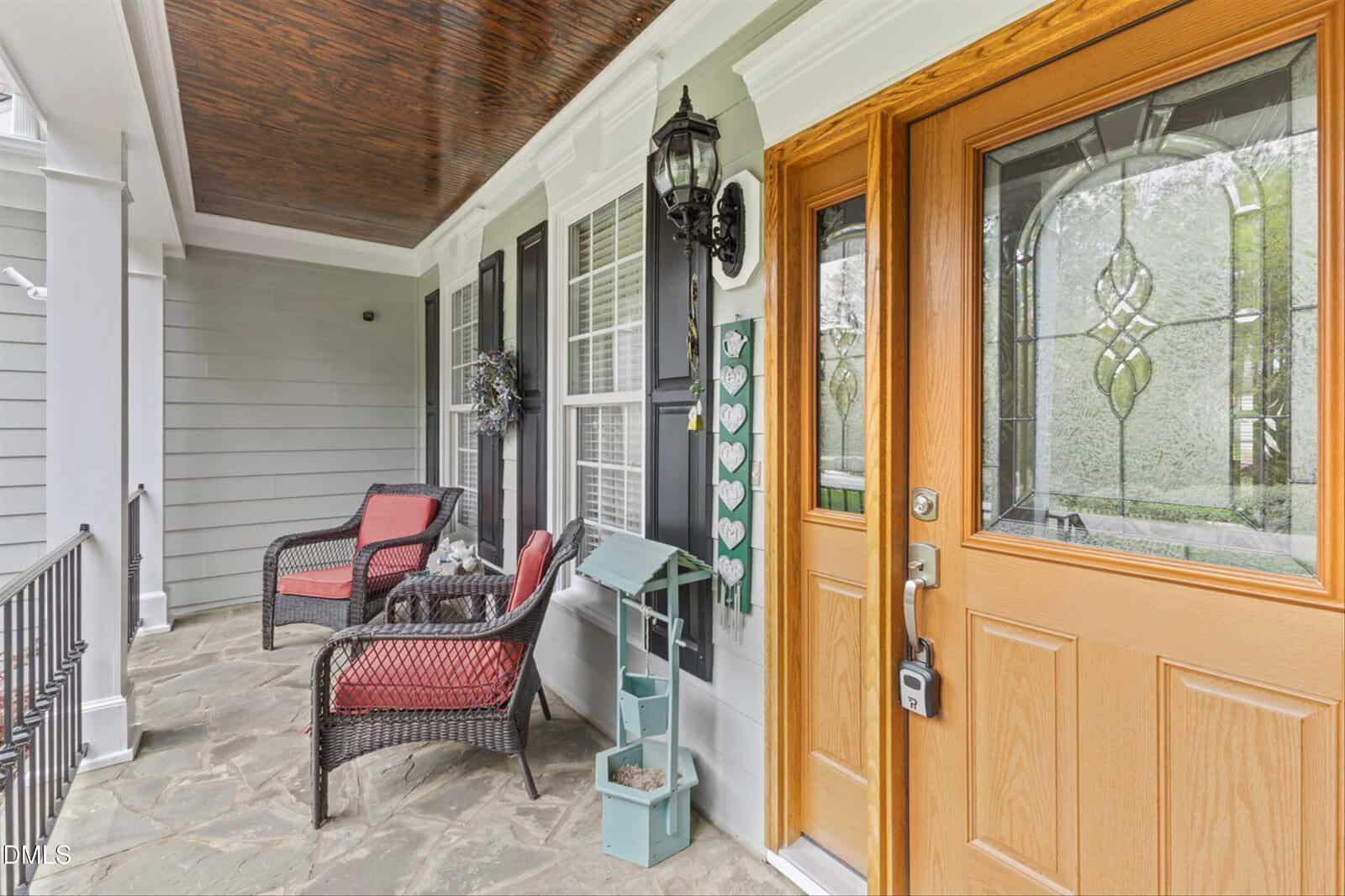 A front porch with stone flooring, wicker chairs with red cushions, a small blue shelf with potted plants, a lantern-style light, and a wooden door with decorative glass. White siding and multiple windows are visible.