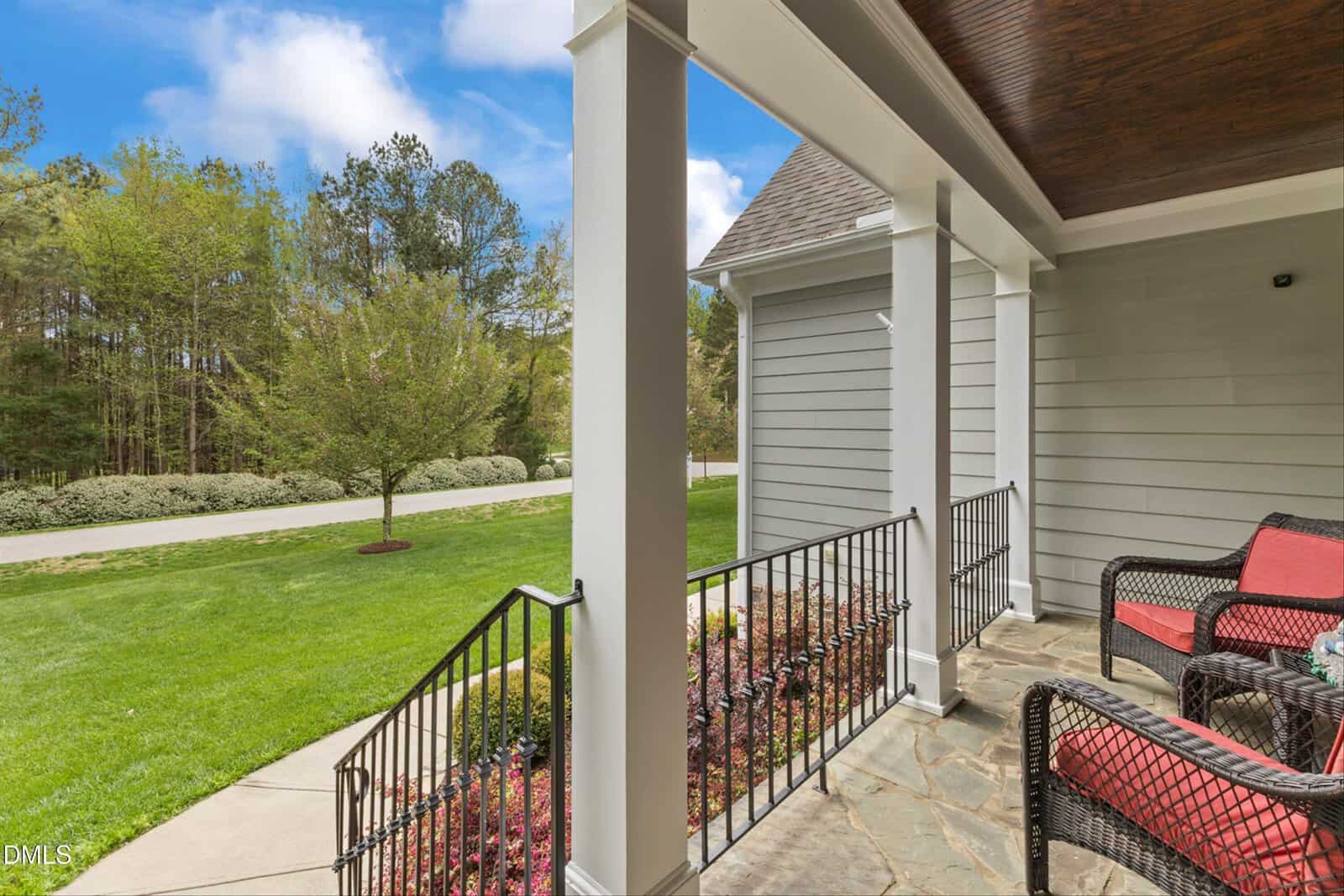 A view from a covered porch with stone flooring, black metal railings, and red cushioned chairs, overlooking a green lawn, sidewalk, and trees in the background under a partly cloudy sky.