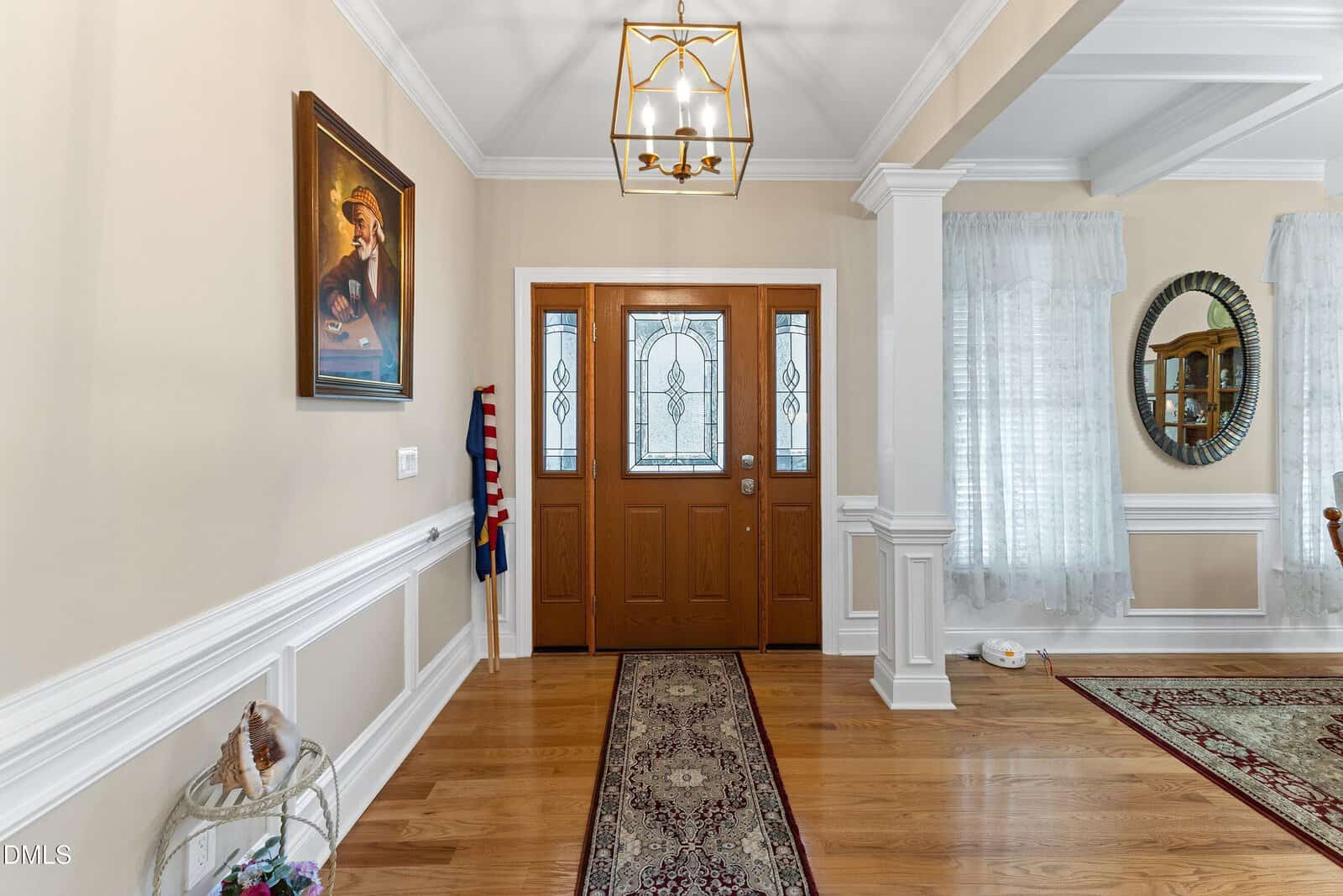 Elegant entryway with double wooden doors, a portrait on the left wall, an American flag, patterned rugs, light hardwood floors, and white wainscoting with crown molding. Large windows with sheer curtains are visible.