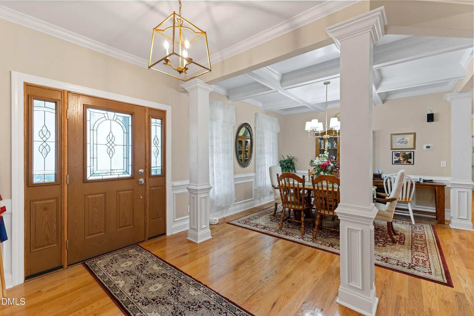 Elegant dining room with hardwood floors, wooden double doors with frosted glass, chandelier lighting, a dining table with flowers, and classic column details dividing the entryway and dining area.