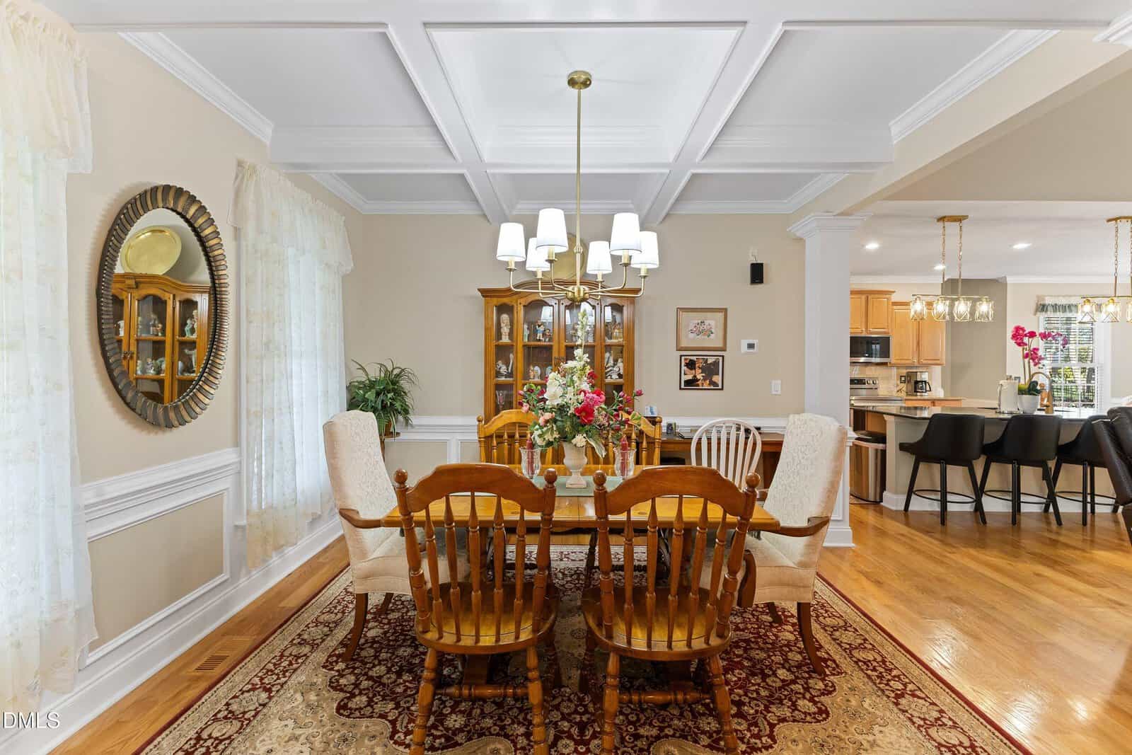 Elegant dining room with wooden table and chairs, floral centerpiece, chandelier above, china cabinet, large mirror, and view into a modern kitchen with barstools. Hardwood floors and decorative rug complete the scene.