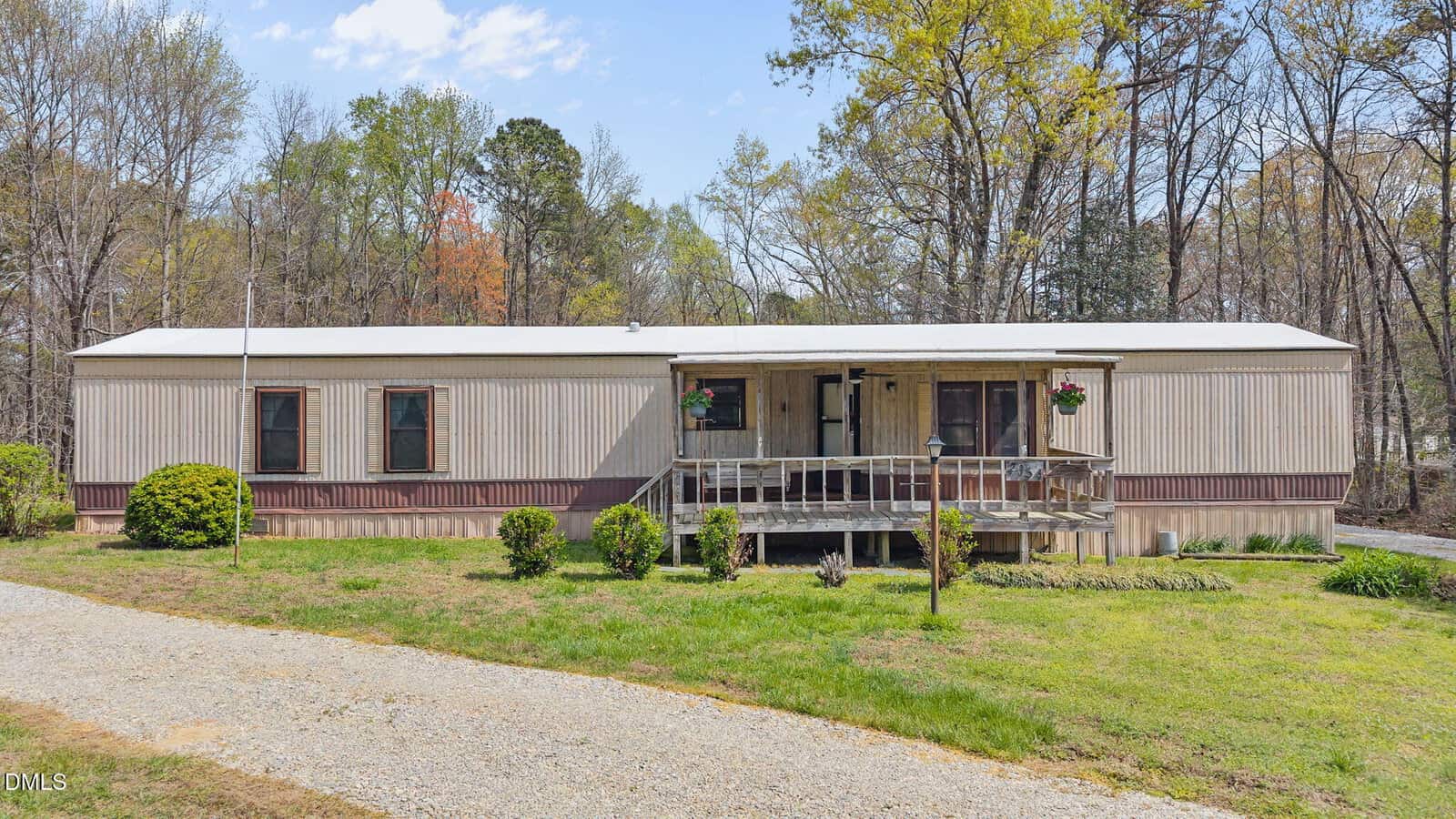 A beige mobile home with a covered front porch and two hanging flower baskets sits on a grassy lot with shrubs, a gravel driveway, and trees in the background.