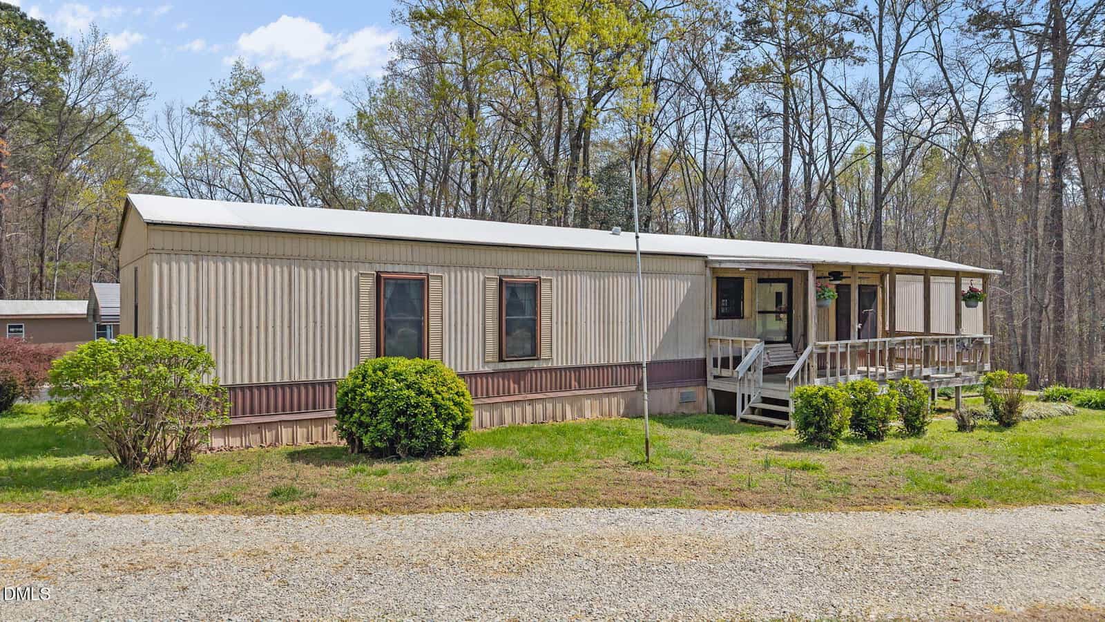 A beige mobile home with brown trim sits on a grassy lot with a gravel driveway. The home has several windows, a small covered front porch with steps, and is surrounded by bushes and trees in the background.