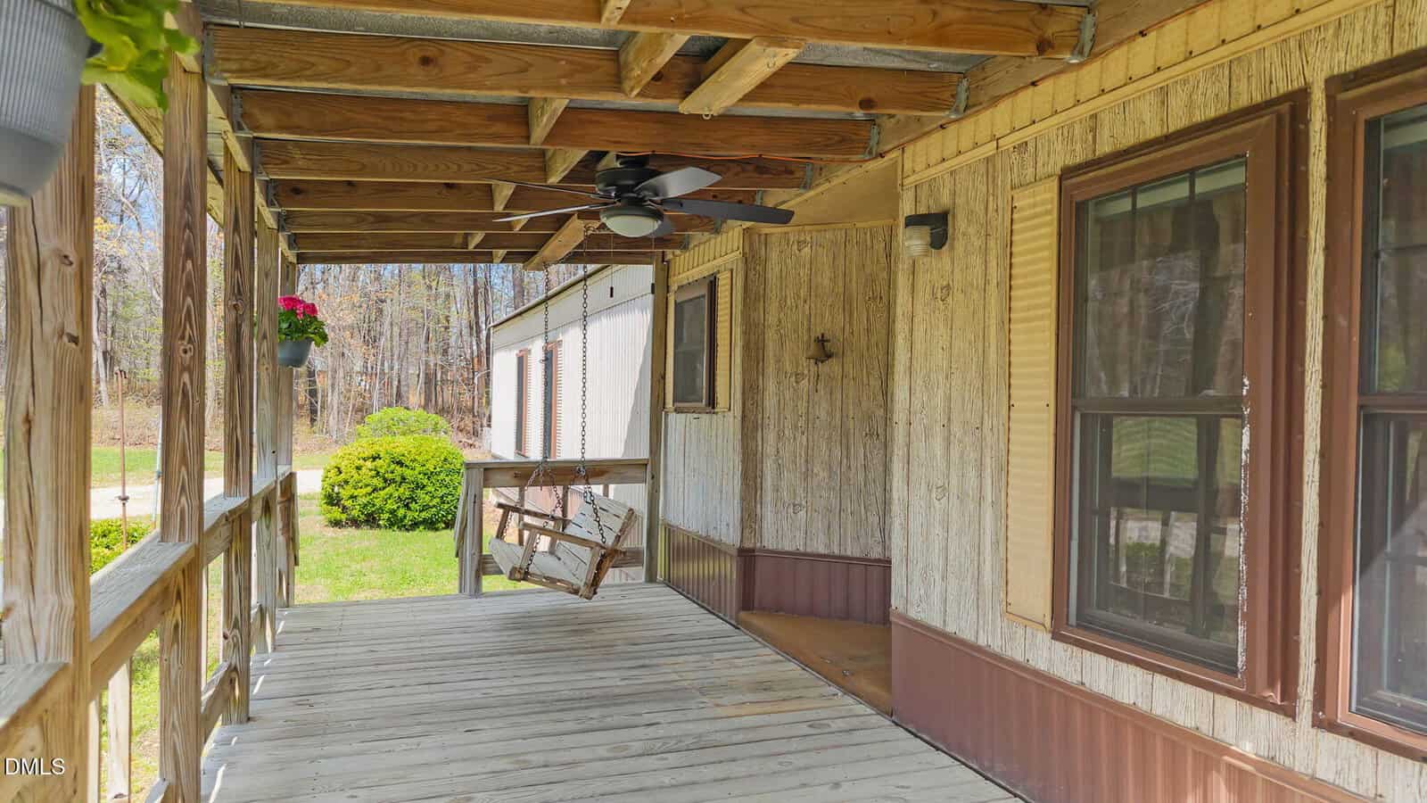 A wooden porch with a ceiling fan, a wooden swing, potted flowers, and railings. The porch is attached to a beige house with brown trim and overlooks a grassy yard with bushes and trees in the background.