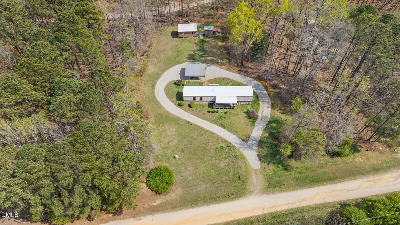 Aerial view of a single-story house with a gray roof, a circular gravel driveway, a small outbuilding, and a larger shed, surrounded by trees and grassy areas near a dirt road.
