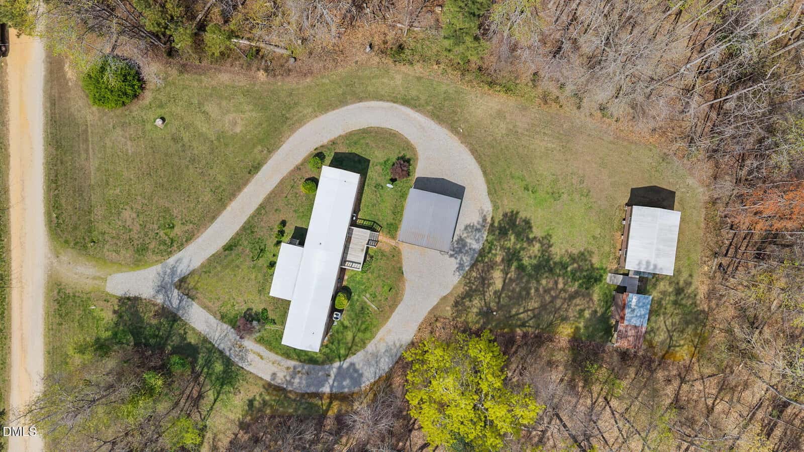 Aerial view of a property with a long white building, a curved gravel driveway, a covered parking area, a smaller outbuilding, and surrounding lawn and trees.