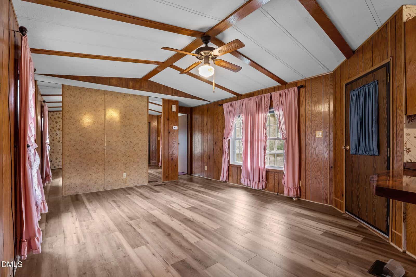 A spacious room with wood panel walls, light wood floors, pink curtains on windows, a ceiling fan with a light, and visible doorways leading to adjoining rooms.