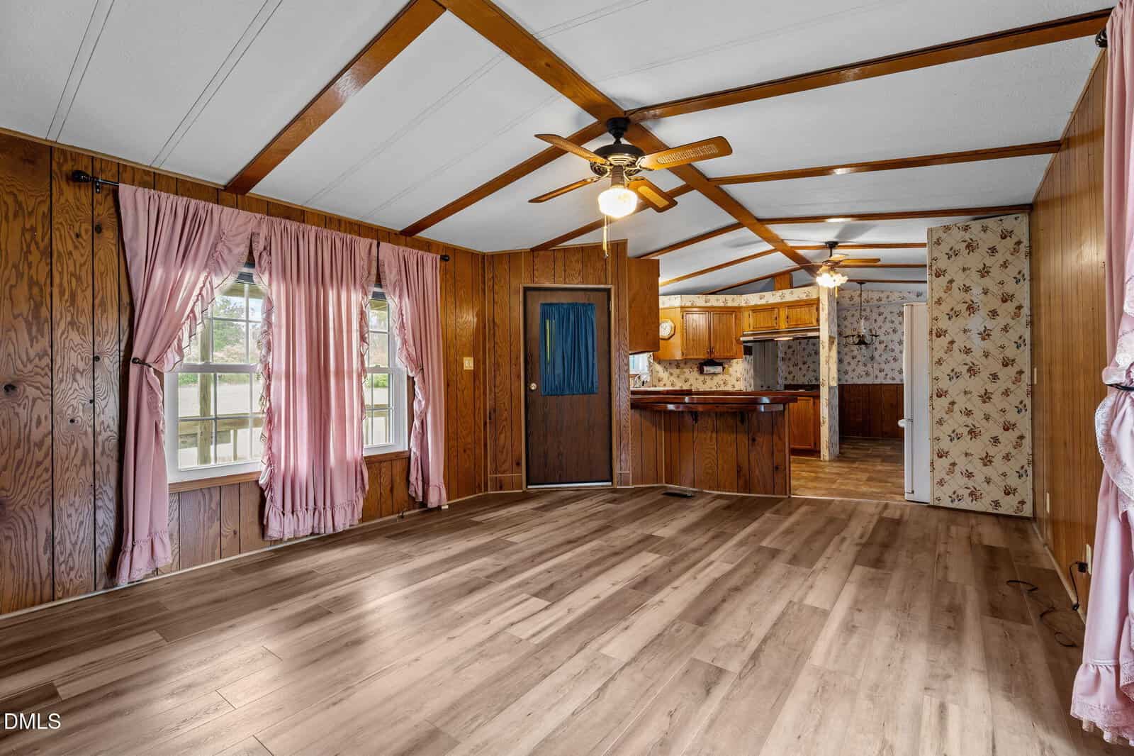 A spacious room with wood paneling, light wood floors, two ceiling fans, pink curtains on windows, and a view into a kitchen with floral wallpaper.