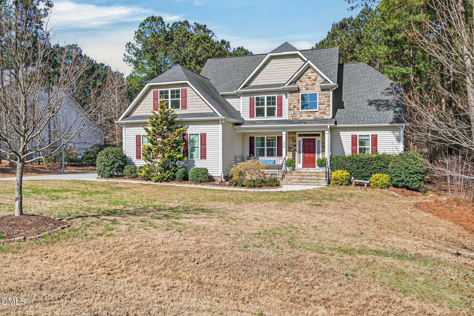 Two-story suburban house with beige siding, red front door, and stone accents. The yard has a few shrubs, a small tree without leaves, and a lawn with brown grass, surrounded by trees and a clear sky overhead.