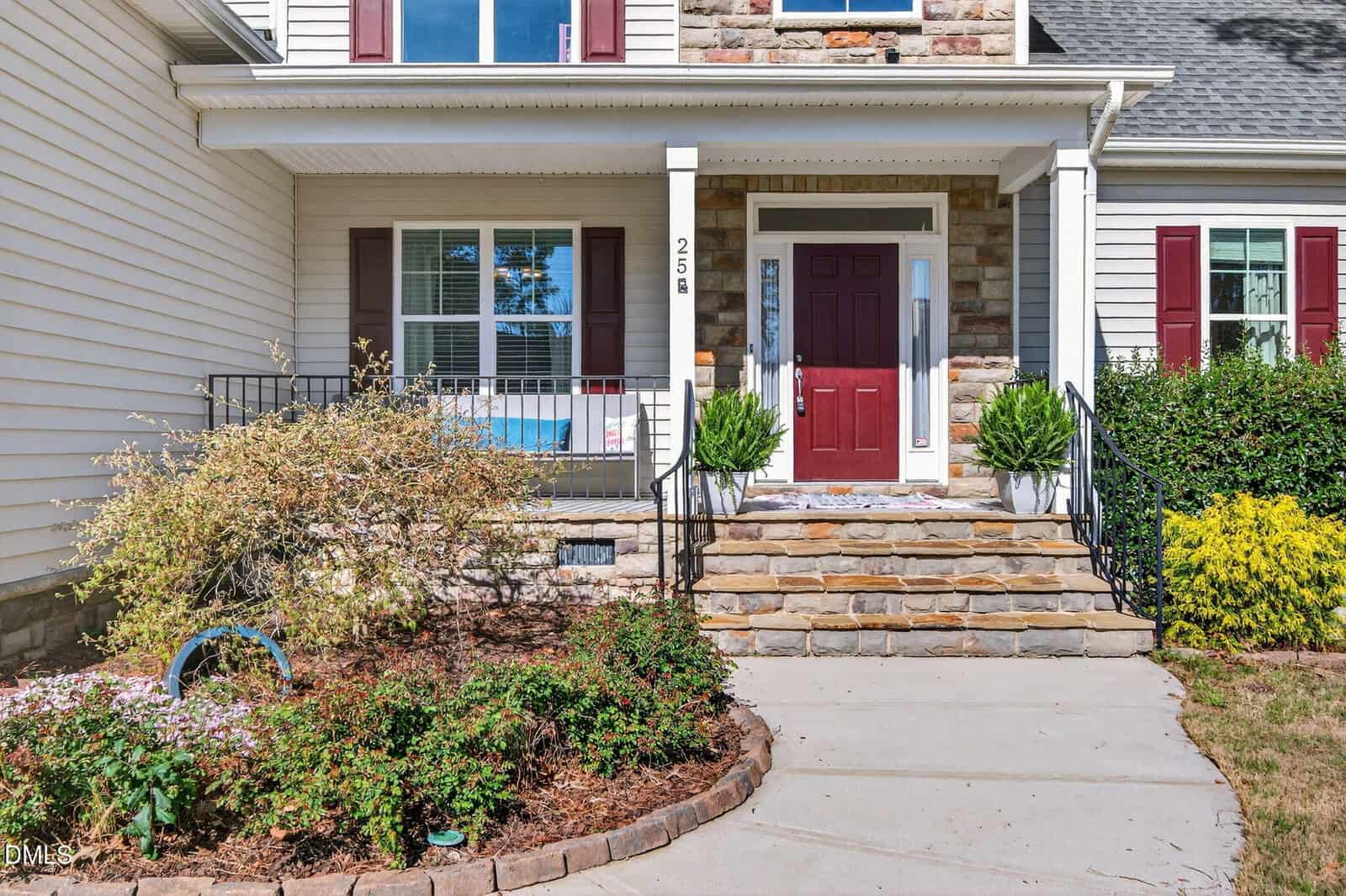 A house entrance with stone steps leading to a red front door, white siding, and brick accents. There are two bushes flanking the steps, a window to the left, and a small front garden with shrubs and flowers.