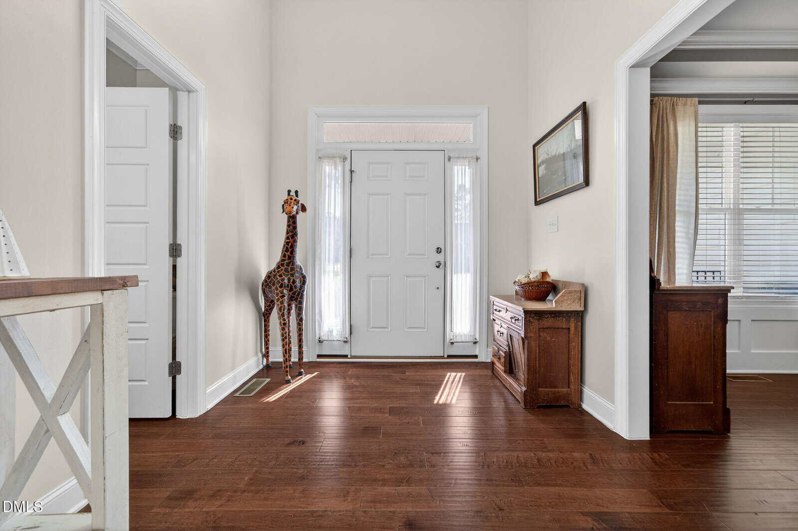 A spacious, well-lit entryway with hardwood floors, a white front door, tall windows with curtains, a decorative giraffe statue, a wooden chest, and framed wall art.
