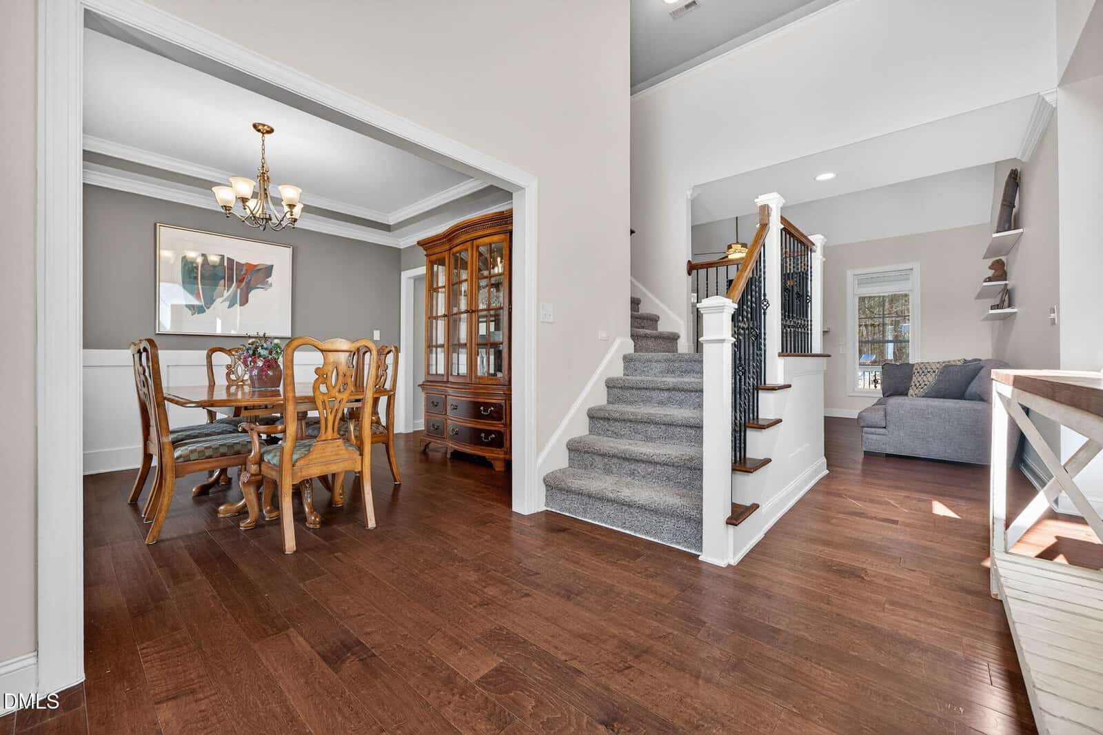 A dining room with a wooden table and chairs is on the left, next to a staircase with carpeted steps leading up. Hardwood floors run throughout, and a hallway with a gray sofa is visible in the background.