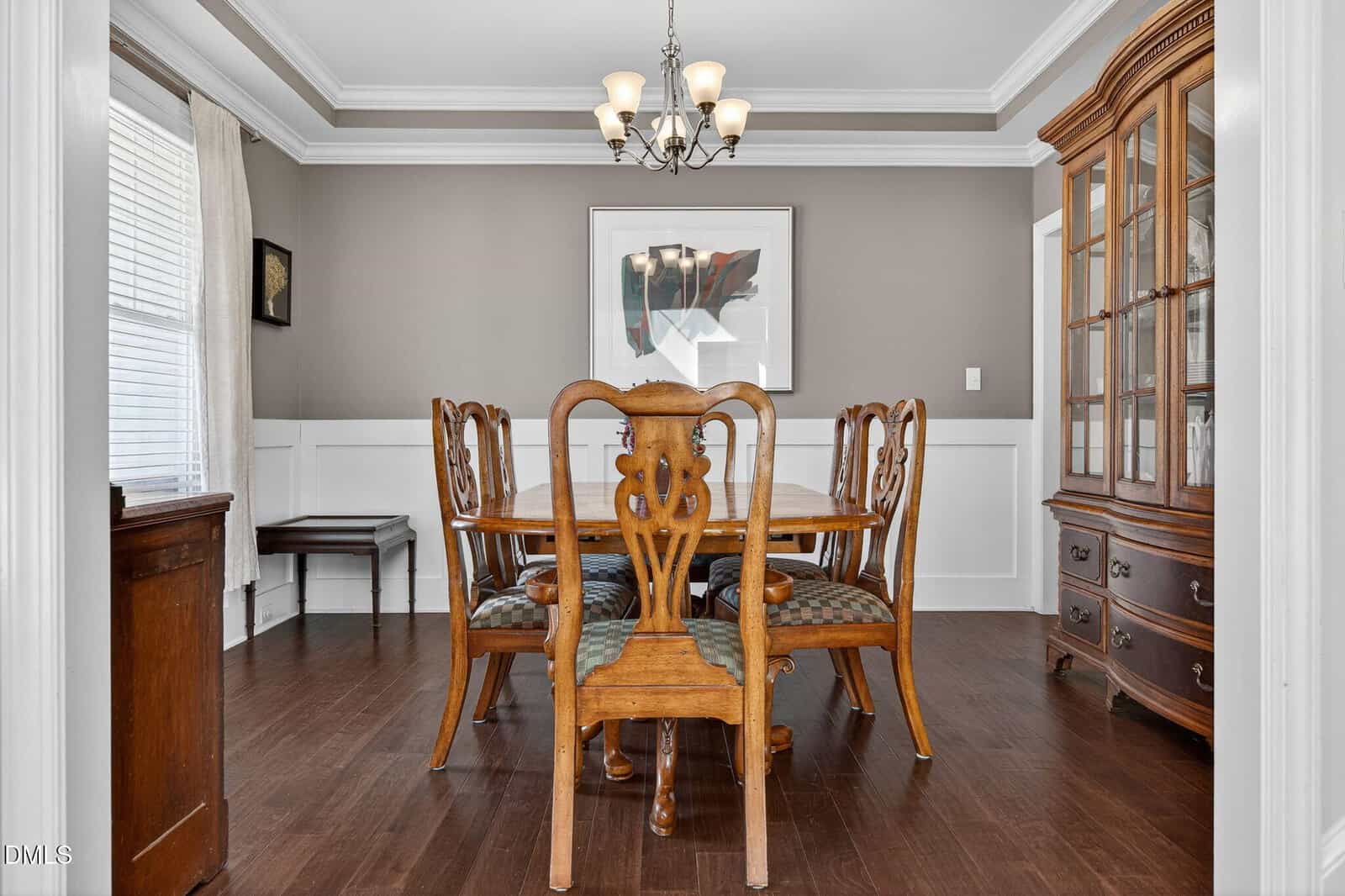 A traditional dining room with a wooden table and six chairs, a chandelier, a glass-front china cabinet, and a framed abstract artwork on the wall. The room has white trim and hardwood floors.